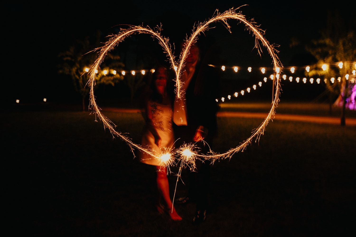 Person drawing a heart shape with a sparkler in the dark with string lights in background.
