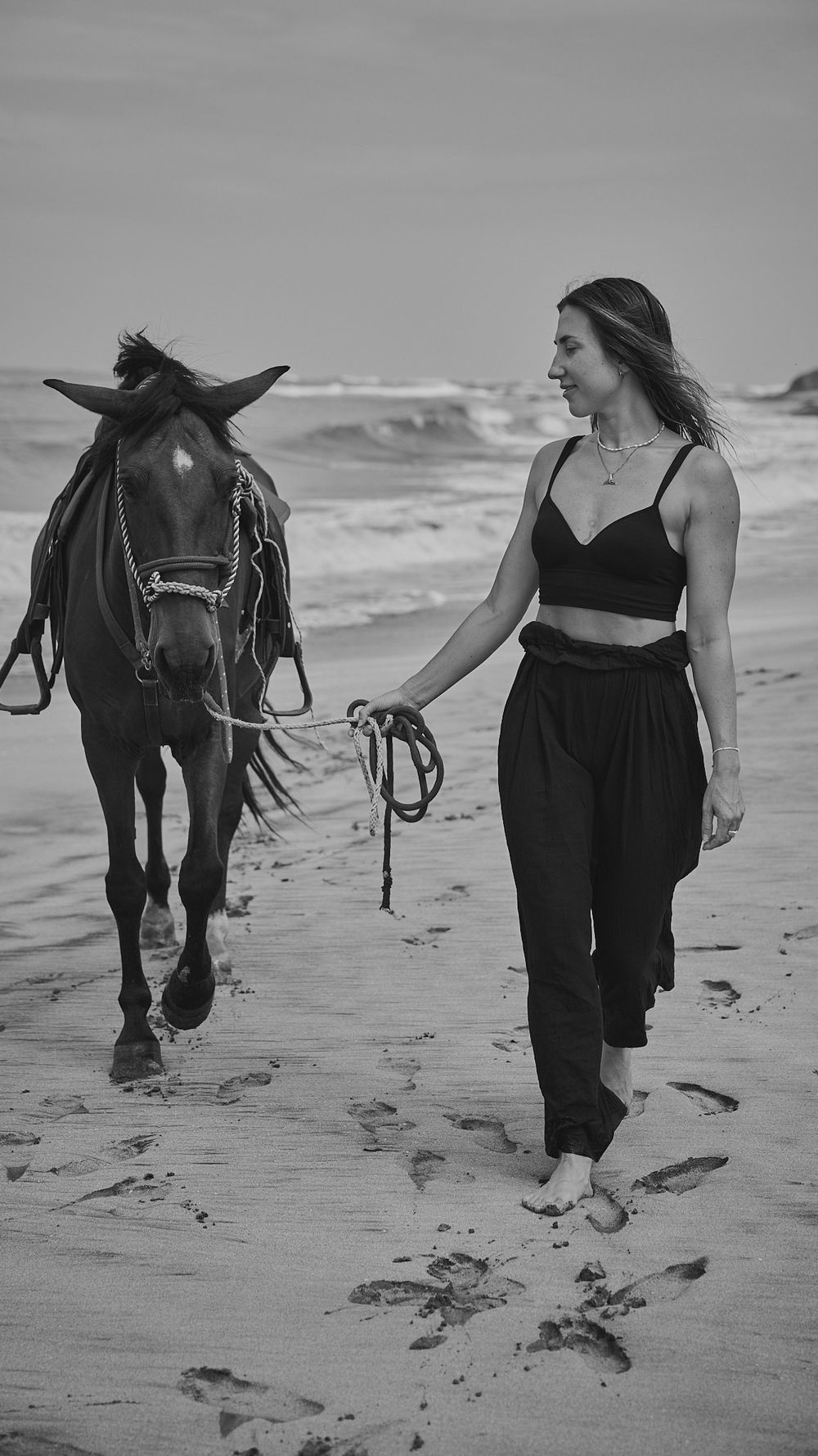A black and white series of images showing a horse and someone walking along a sandy beach near the ocean waves.