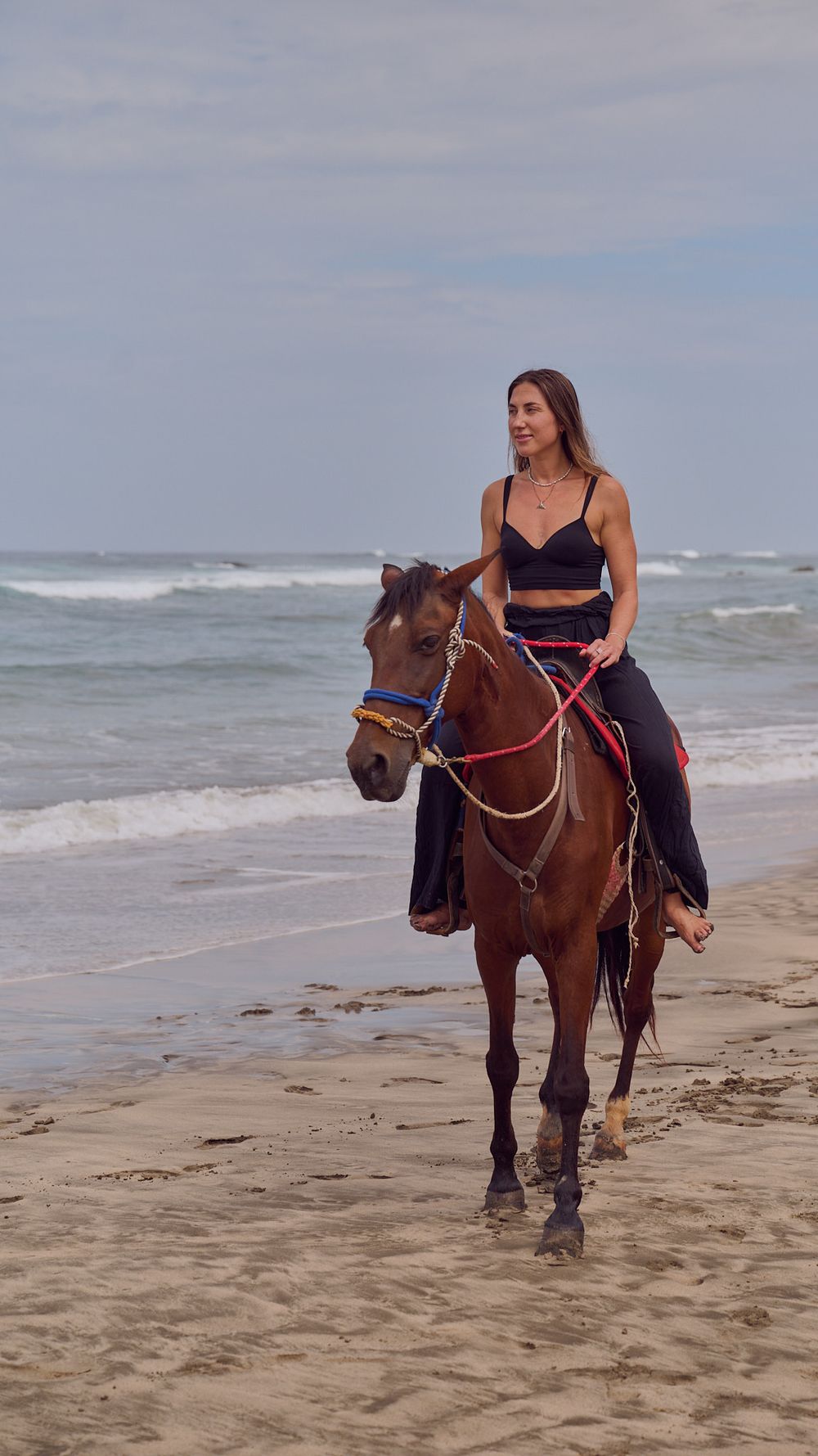 A woman in a navy dress rides a brown horse along the sandy beach with waves in the background.