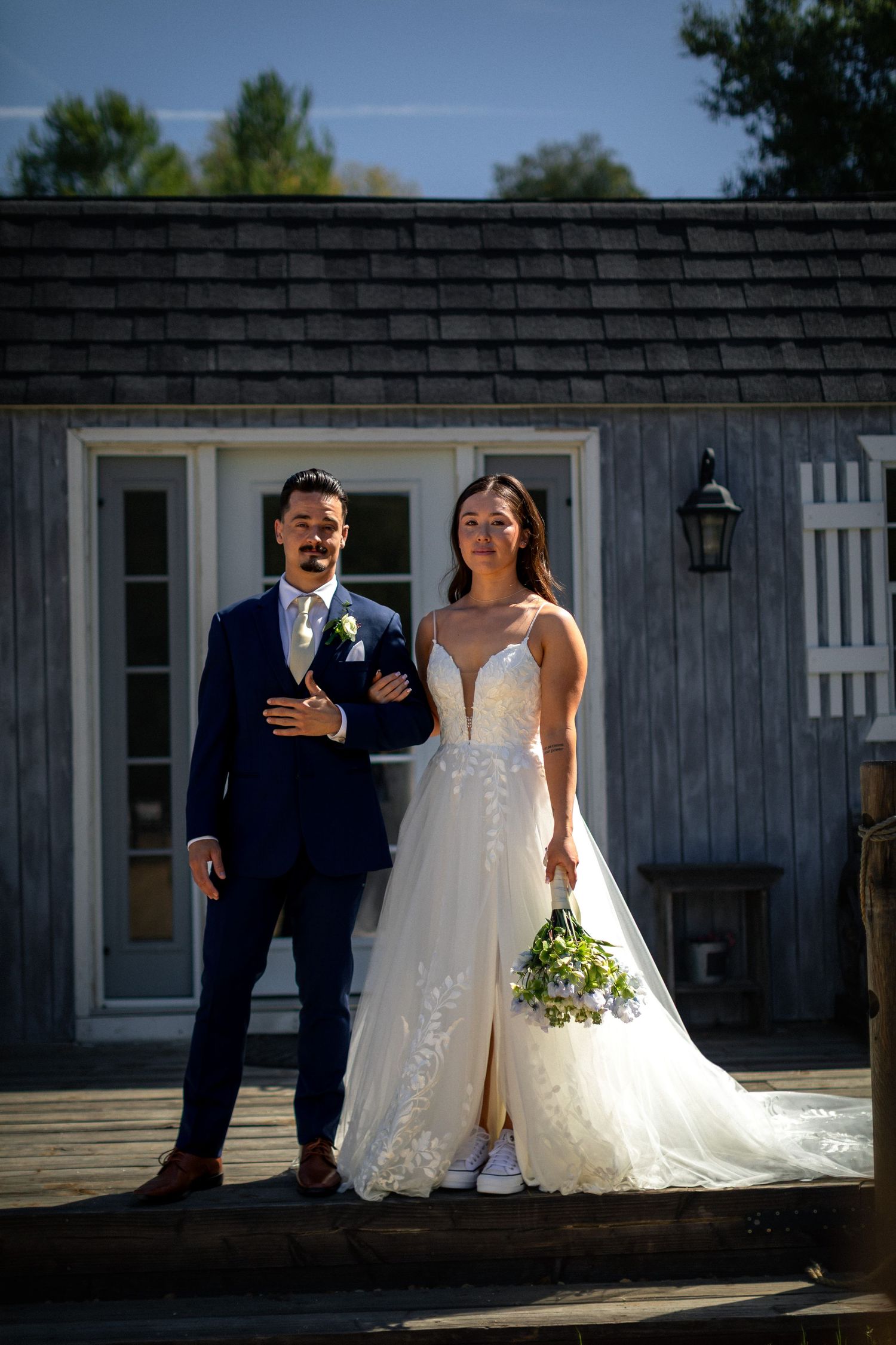 A couple in wedding attire poses in front of a rustic gray building with white trim.