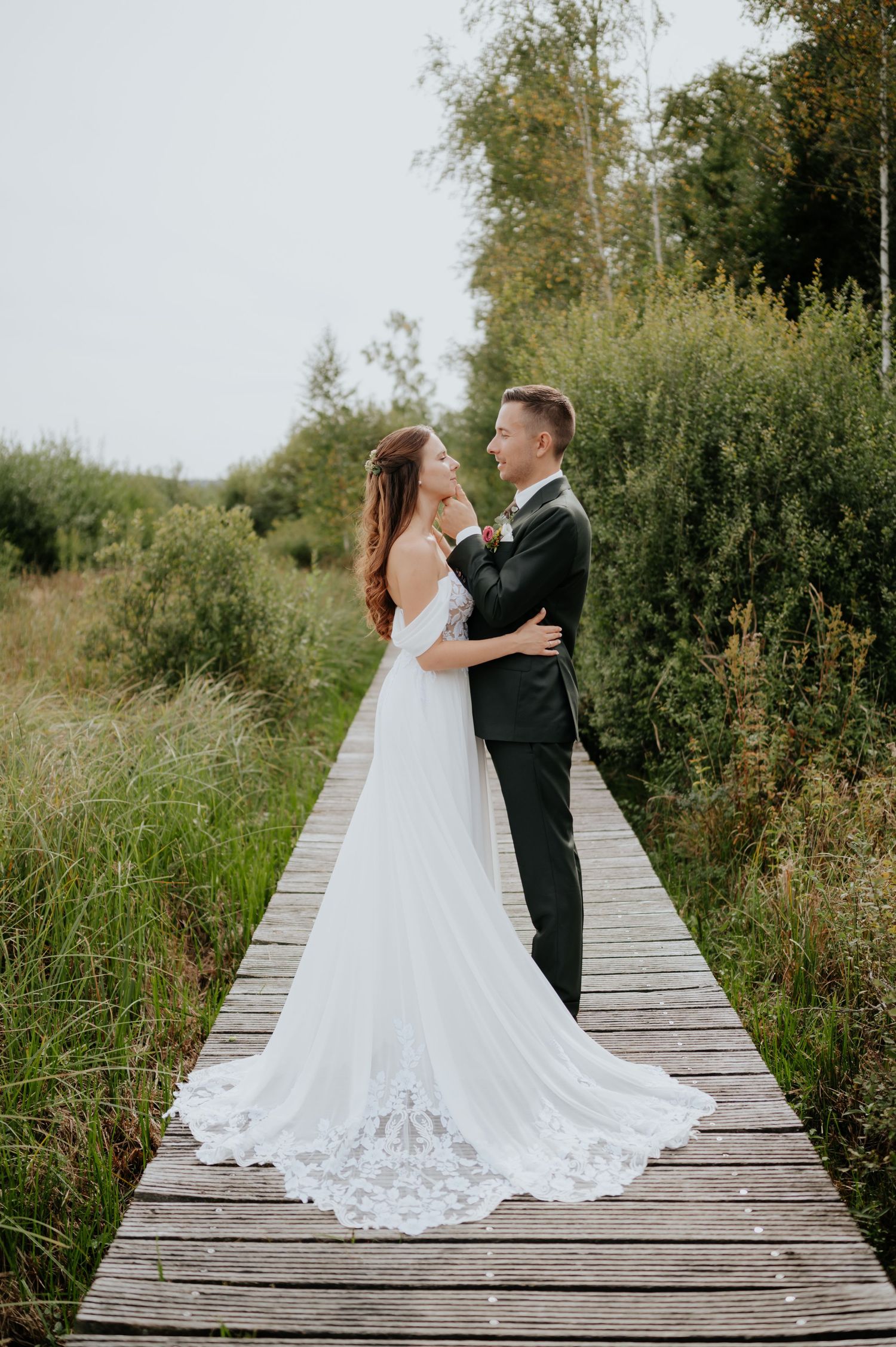 Ein Paar in Hochzeitskleidung genießt intime Momente auf einem Holzsteg, umgeben von hohem Gras und natürlicher Landschaft.