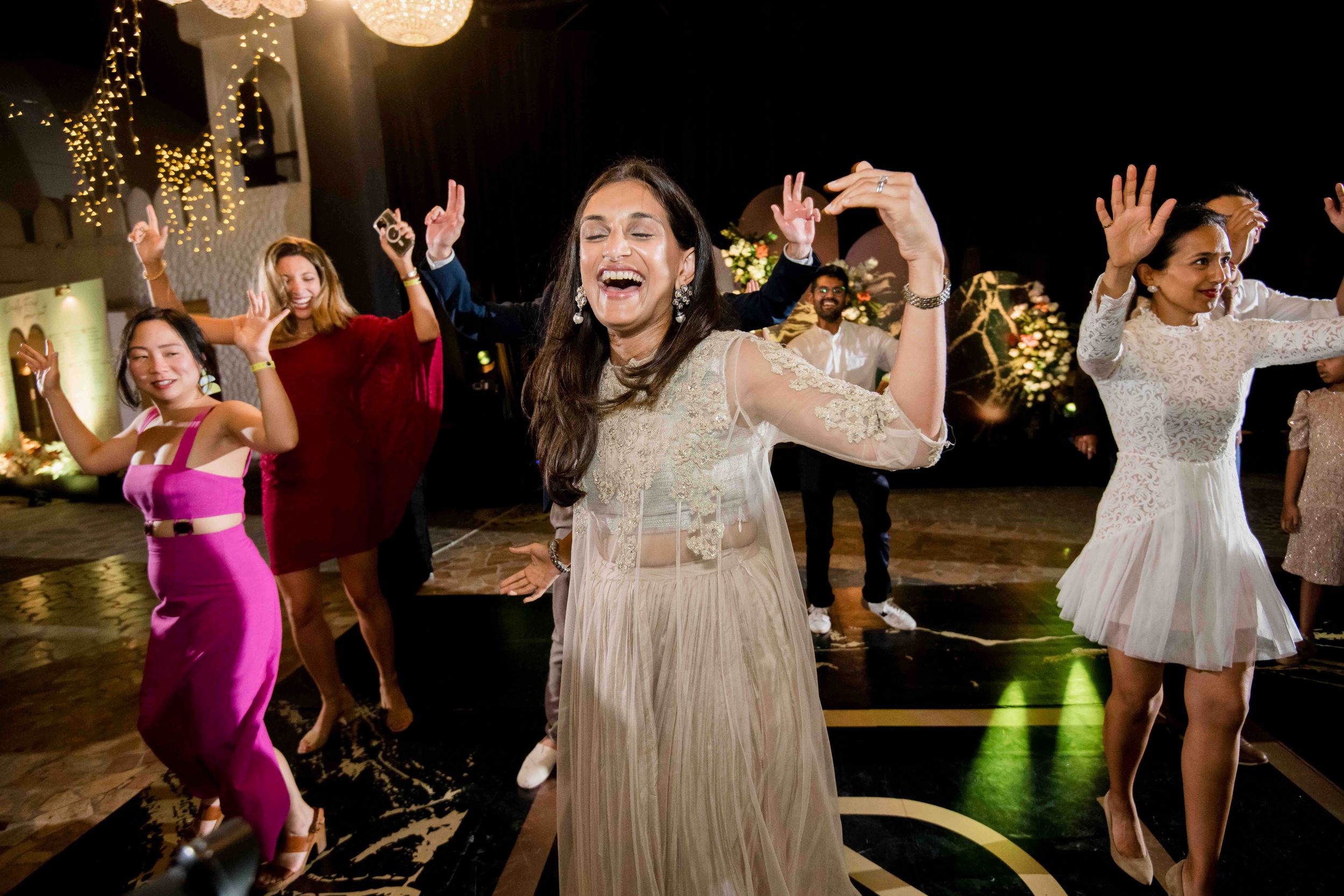 Group of wedding guests dancing and celebrating on an illuminated dance floor at a nighttime reception.