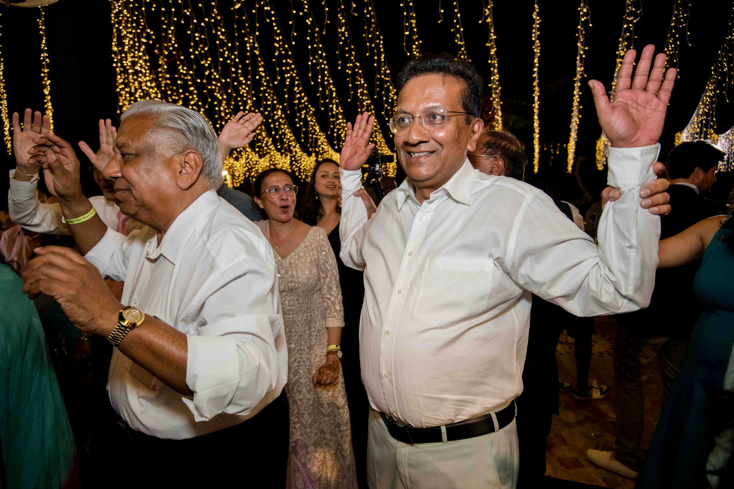 Two people in white shirts dancing and waving their hands with twinkling lights in the background at a celebration.