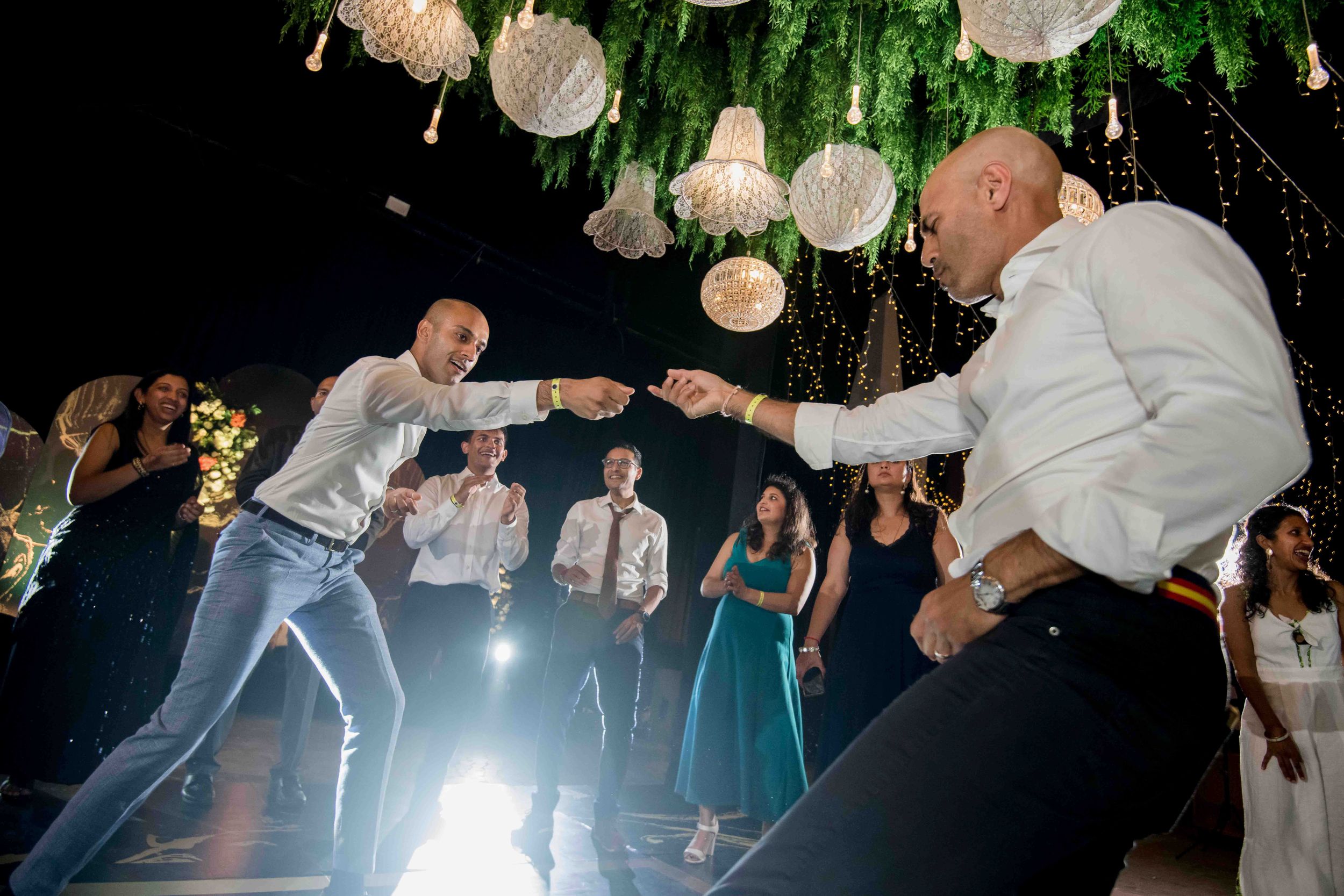 Wedding guests dancing energetically under decorative white lanterns at an evening reception party.