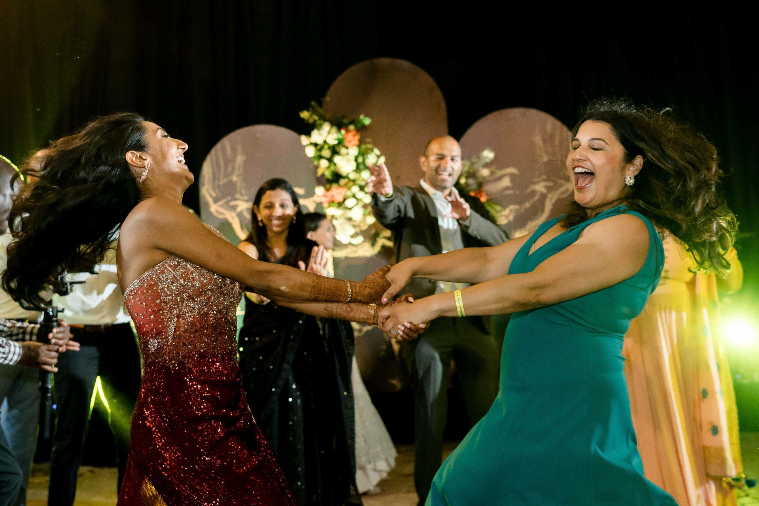 Party guests in red sequin and turquoise dresses dance enthusiastically at an evening reception with glowing lights.