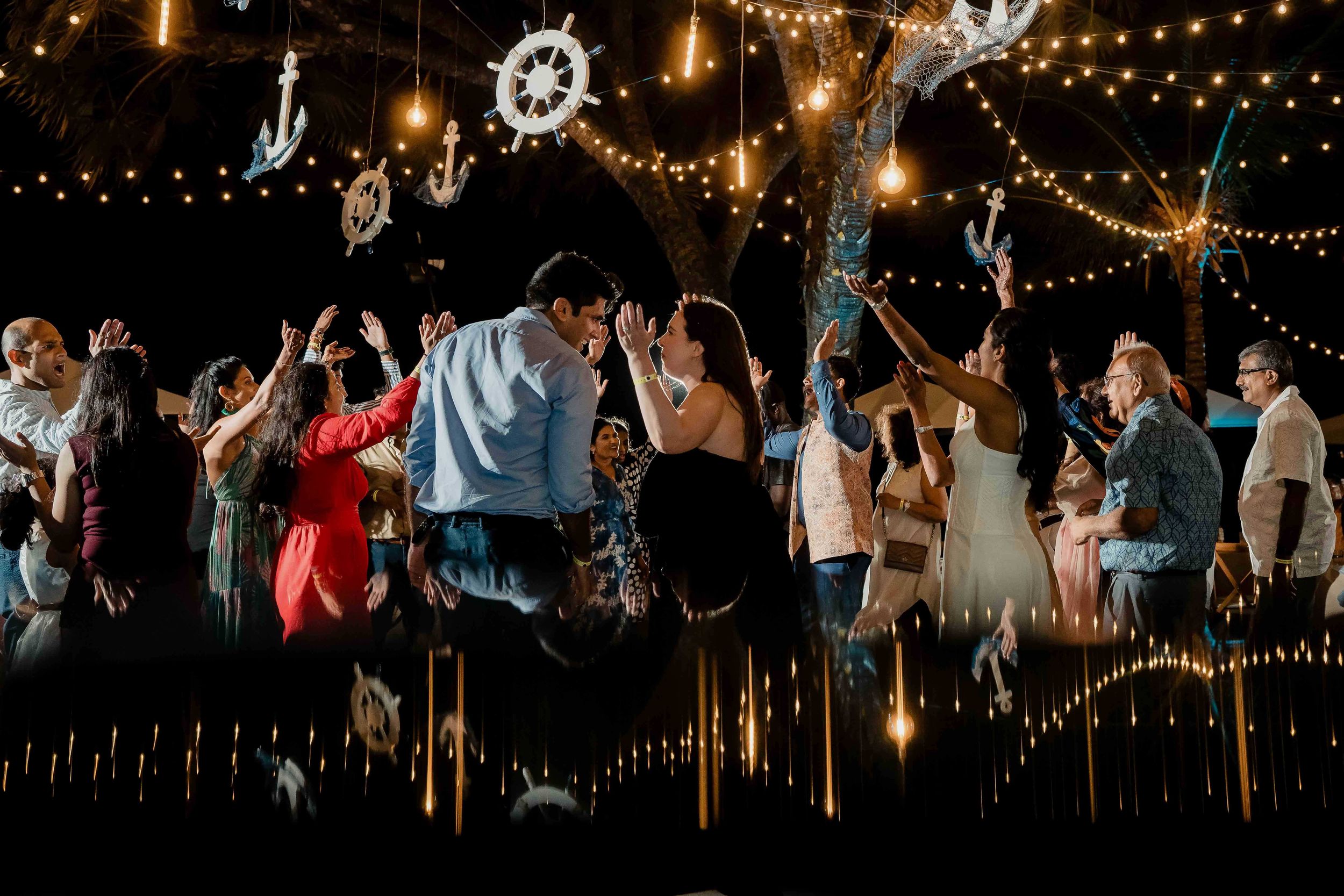 People celebrate and dance under strings of glowing lights at a nighttime outdoor party or reception.