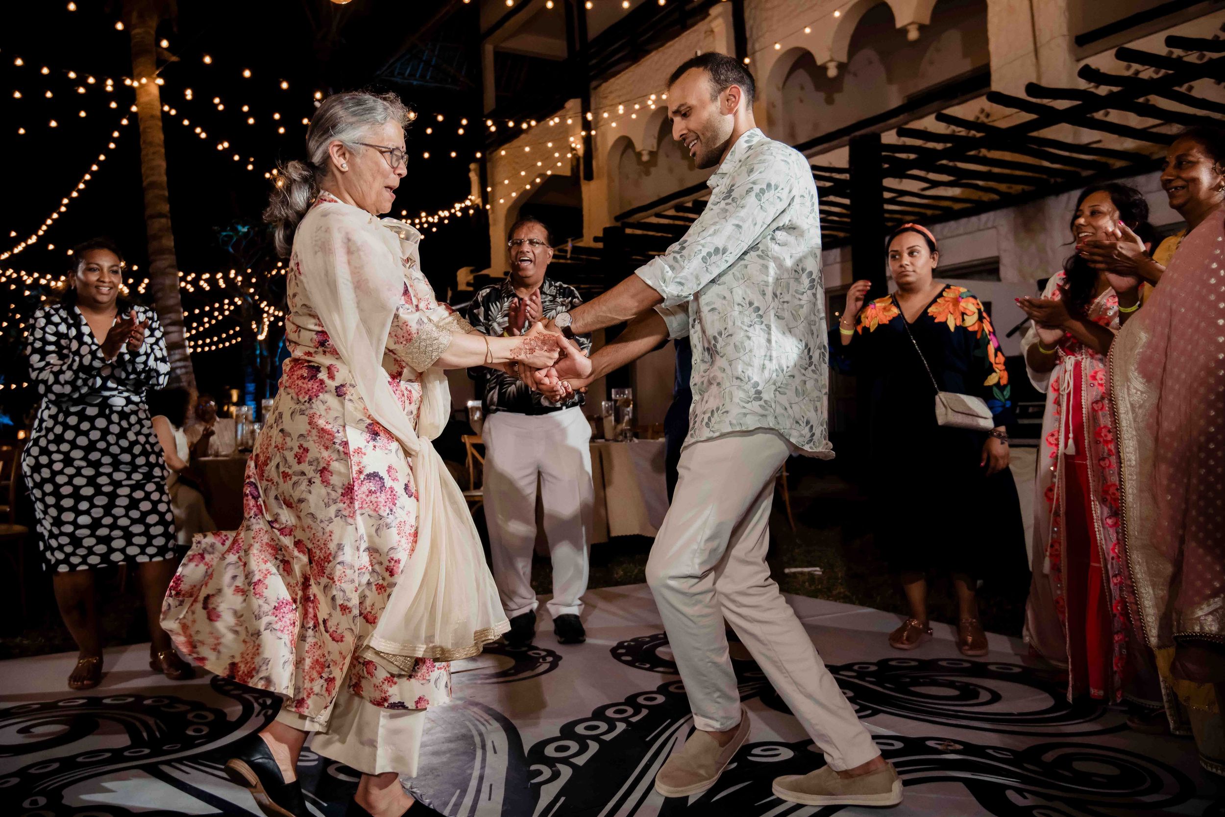 People dance joyfully at a wedding reception under twinkling lights creating a festive evening atmosphere.
