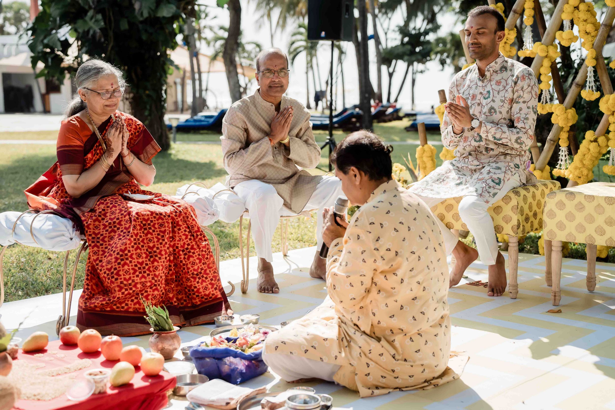 People sit together outdoors participating in a traditional Thai ceremony with offerings and decorations arranged around them.