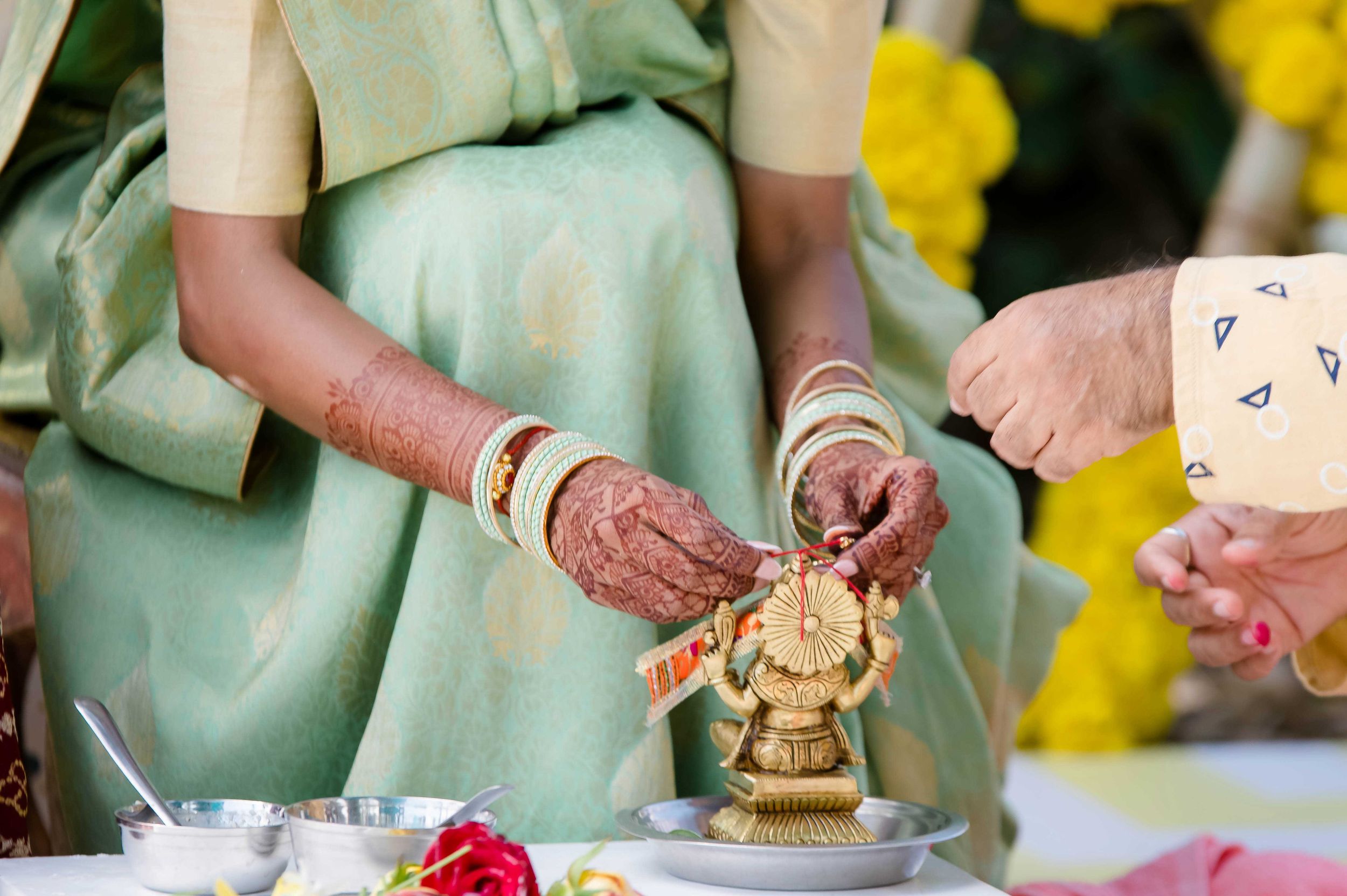 A pair of henna-adorned hands perform a traditional Indian religious ceremony with a small golden statue.