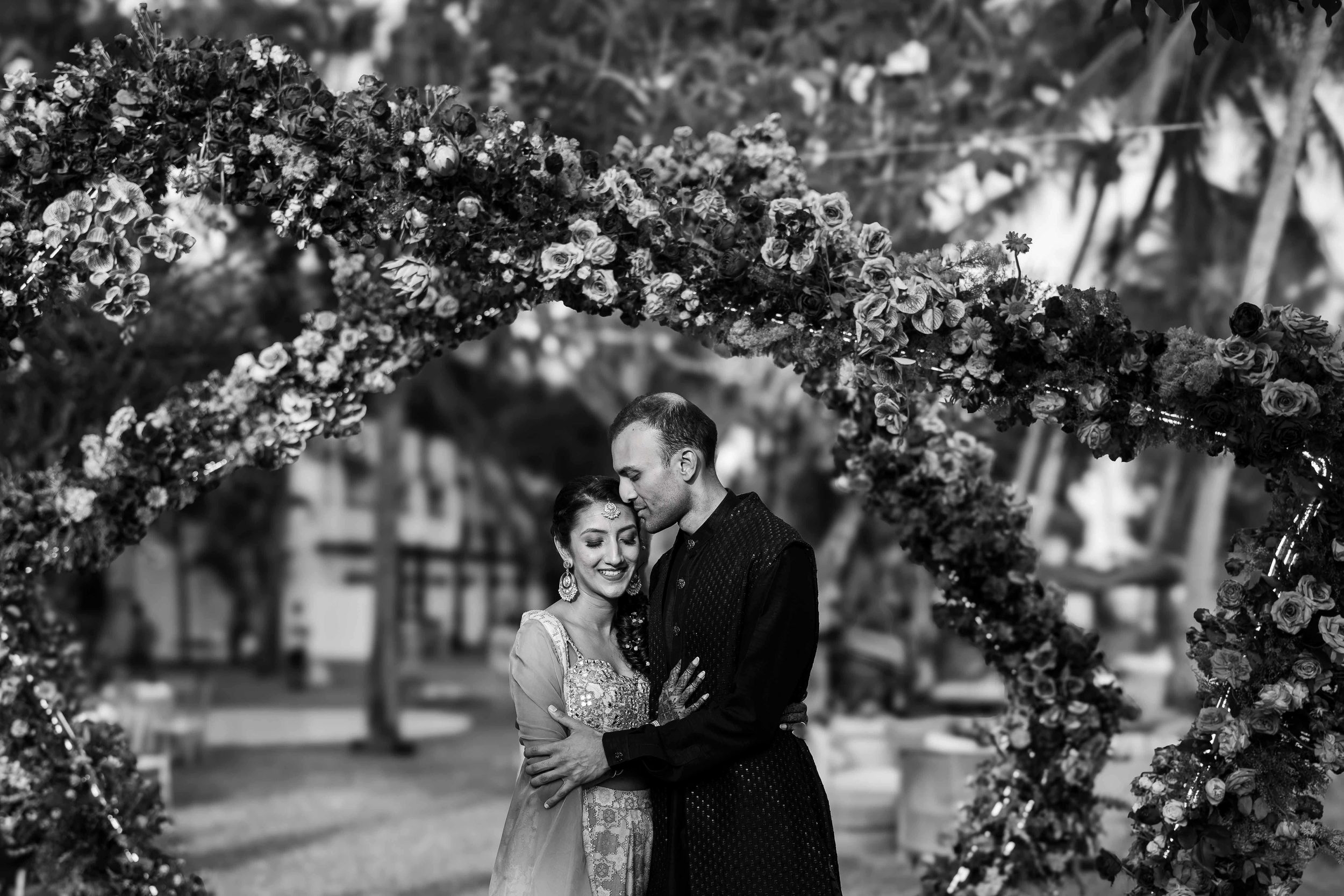 A romantic couple embraces under an elegant floral arch in a serene garden setting.