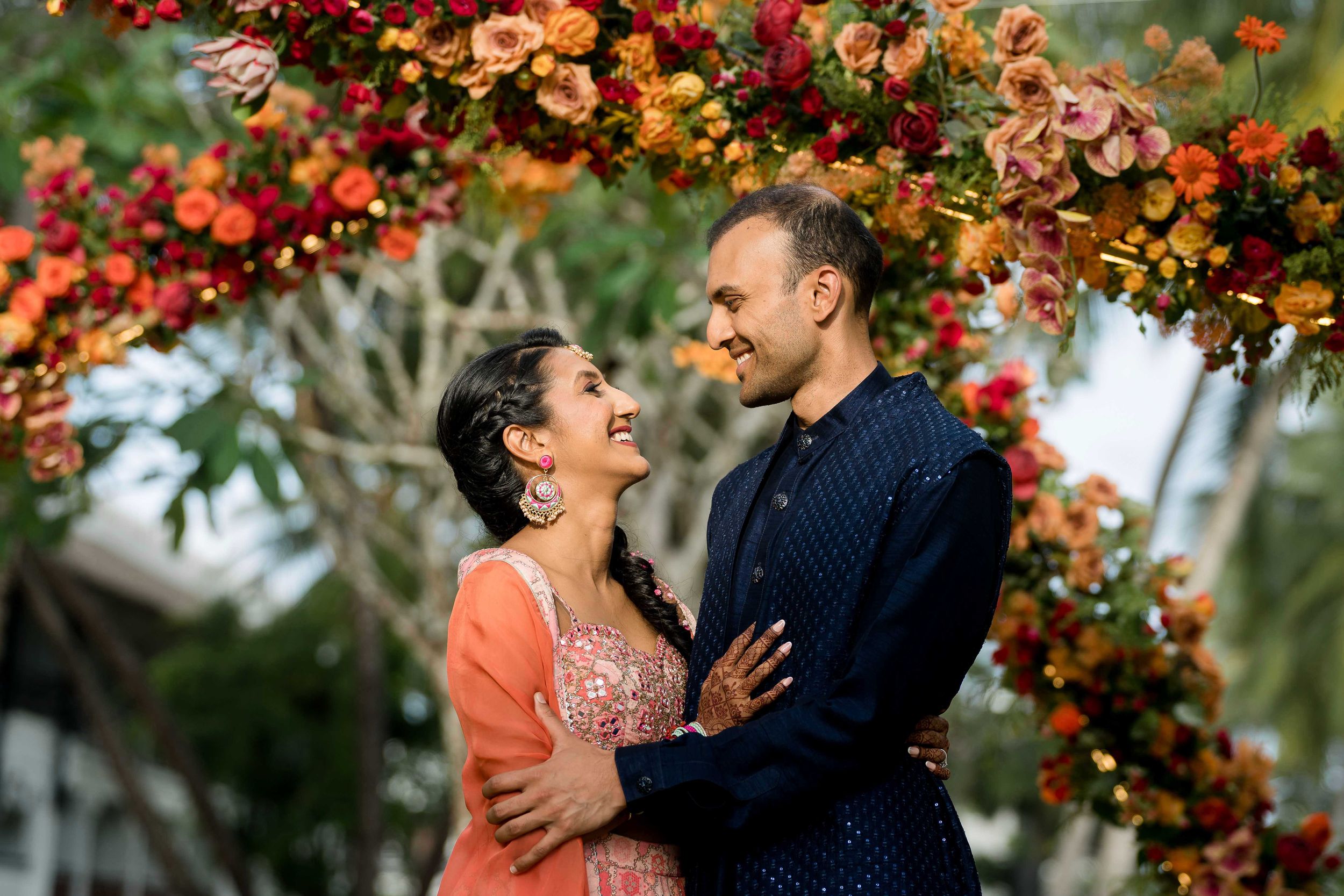 A couple embraces under a floral arch with red and orange blooms against a blurred natural background.