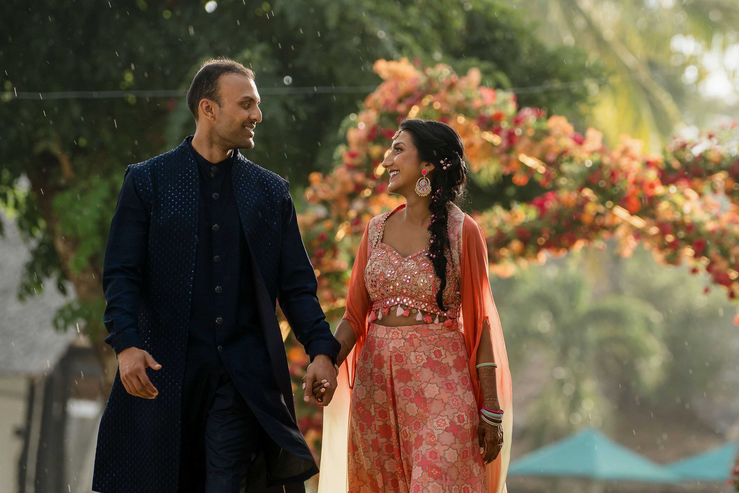 Two people walk together under a tropical archway adorned with vibrant peach-colored flowers in a romantic setting.