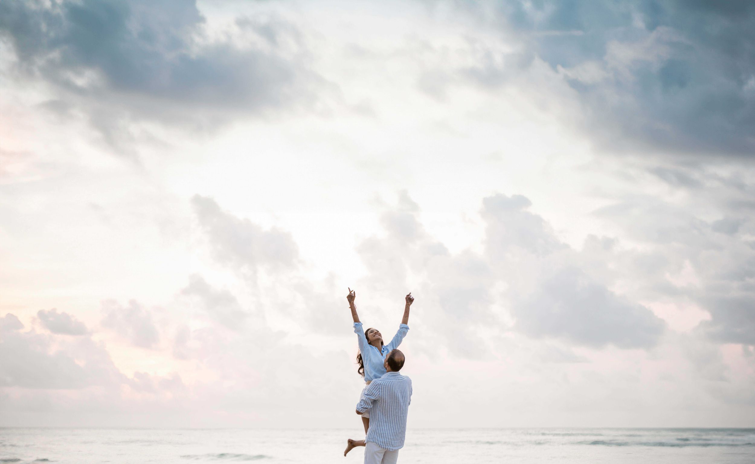 Person celebrating with raised arms in a white shirt against a cloudy beachside panoramic background.
