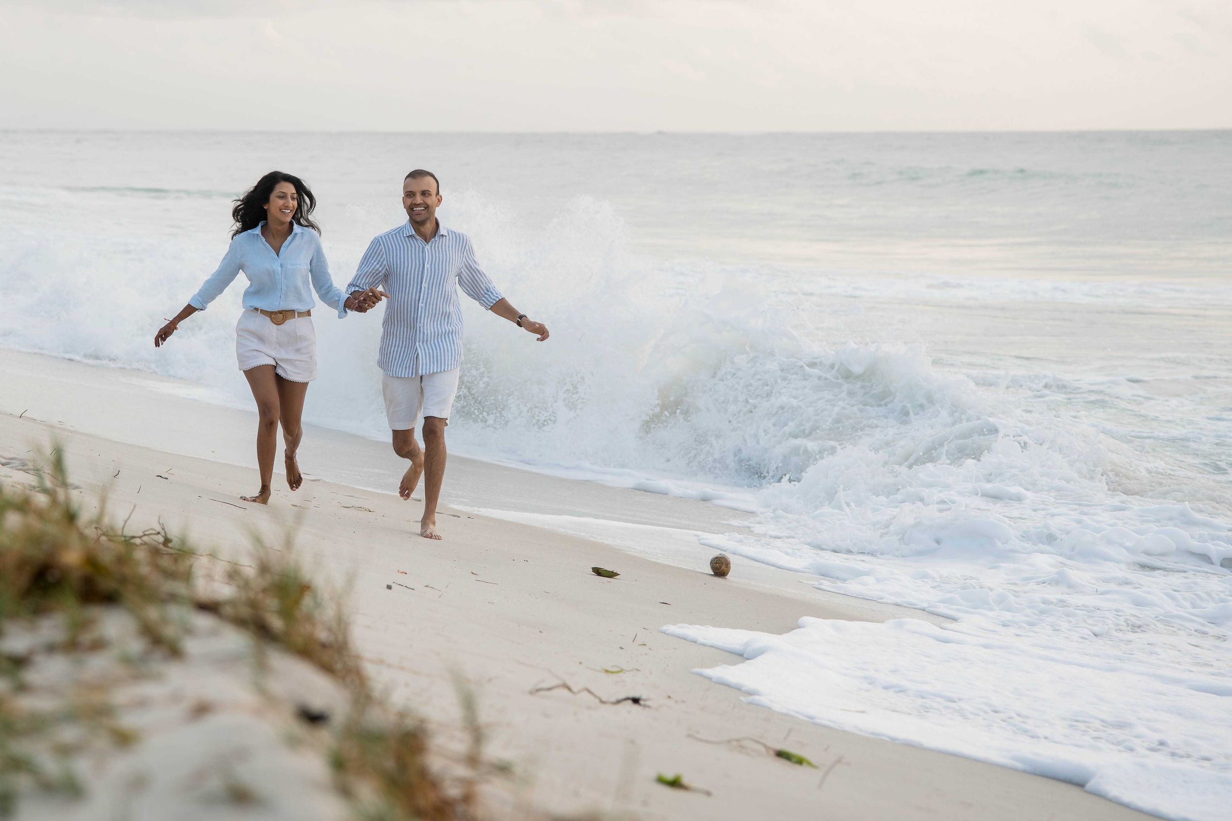 A joyful couple runs together along a sandy beach with crashing waves in the background at sunset.