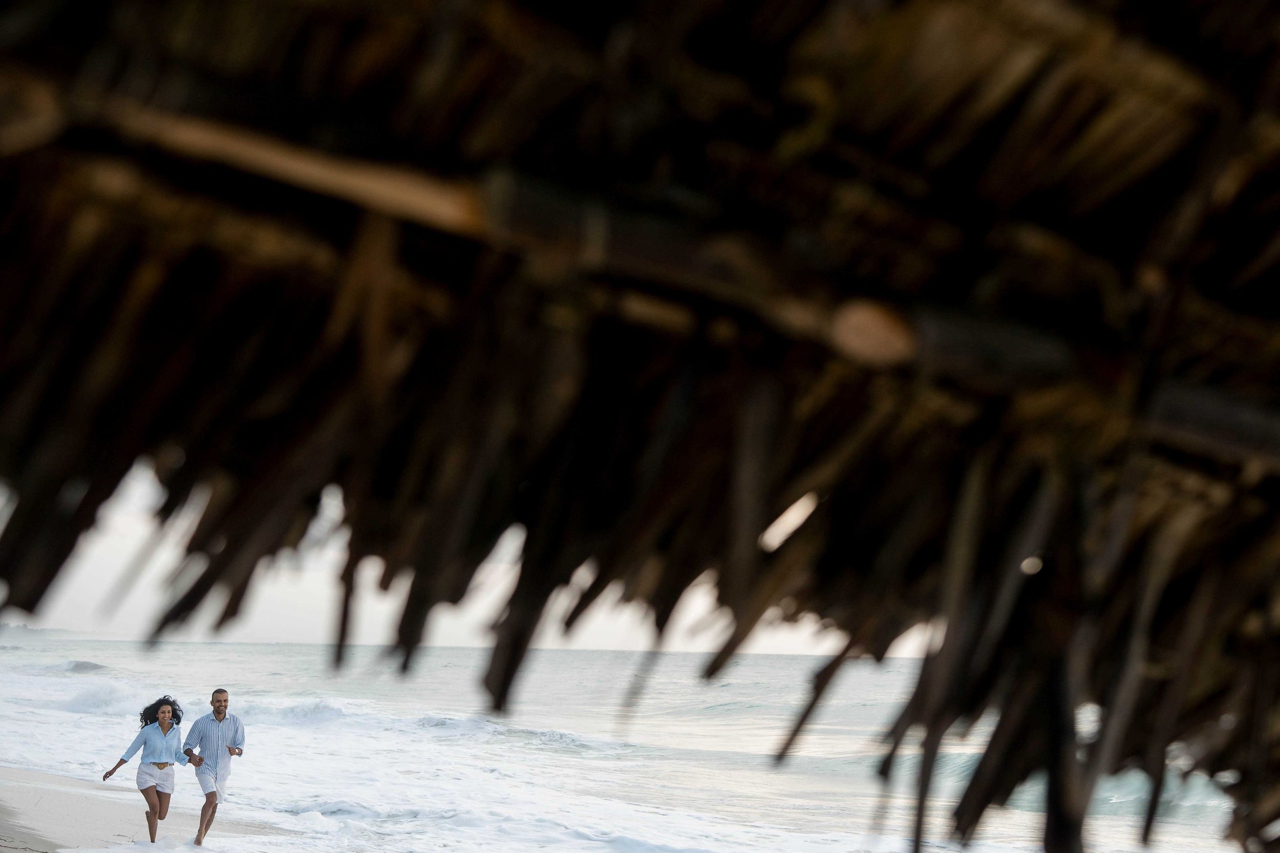 A couple walks beneath a thatched palm roof structure on a tropical beach.