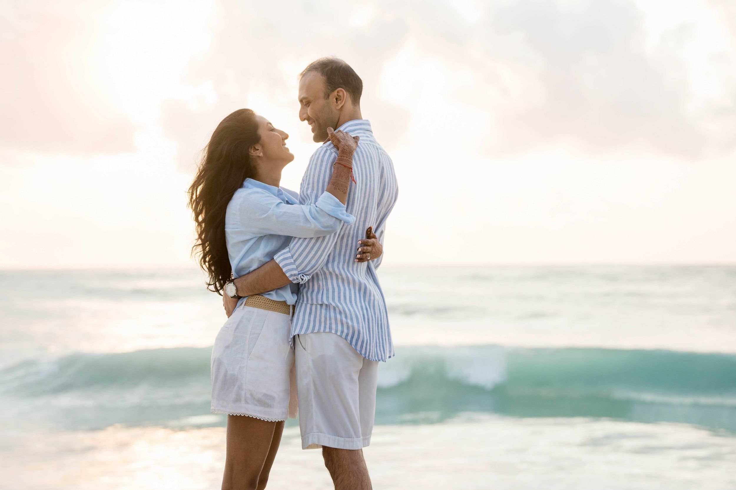 A romantic couple embraces on the beach during sunset with turquoise waves crashing in the background.