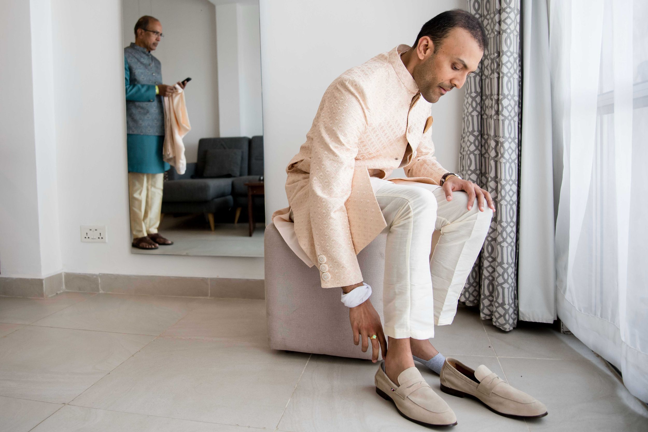 Person in beige sweater puts on shoes near windowsill in bright white modern interior.