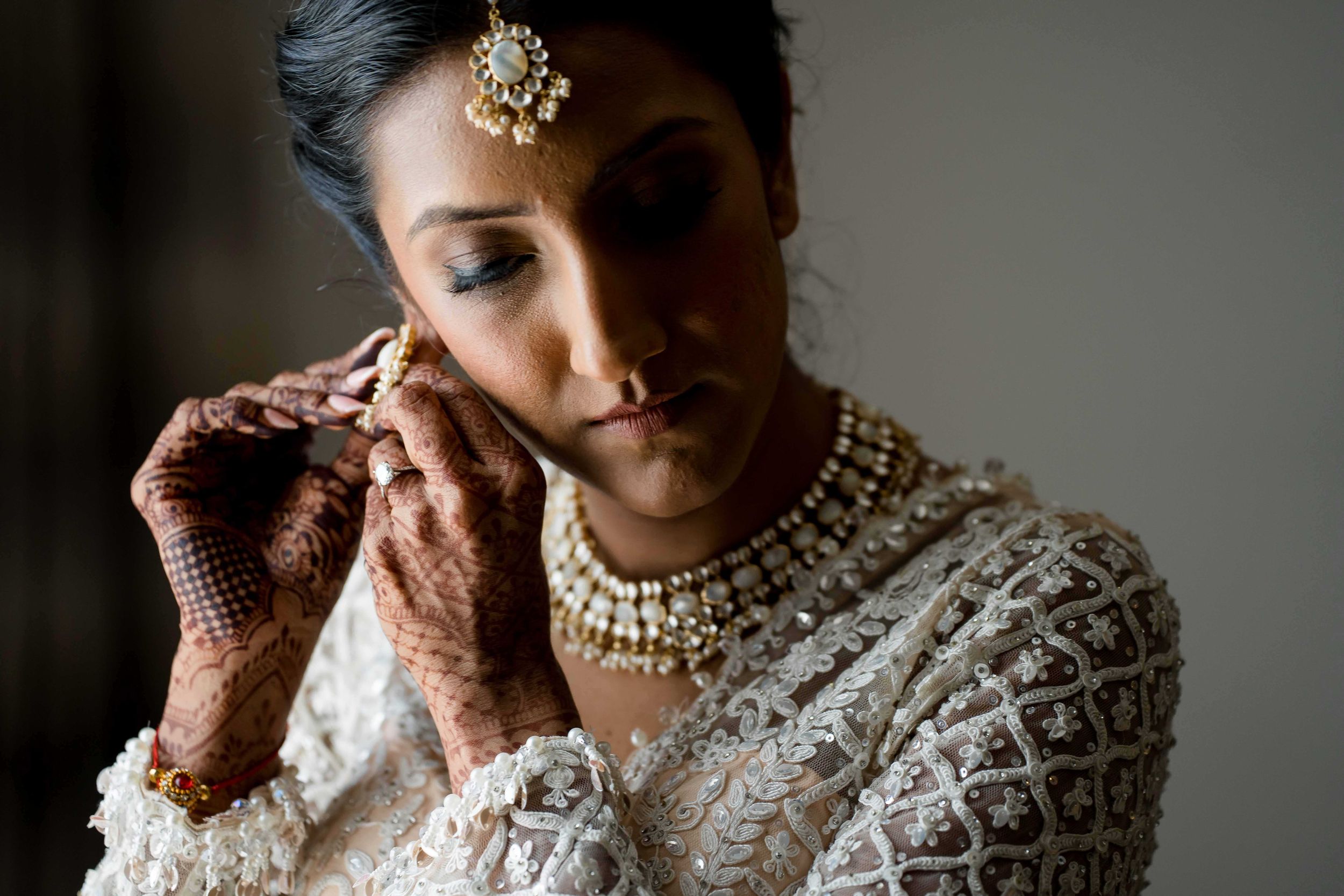 Someone wearing traditional Indian bridal jewelry and white embroidered attire adjusts earrings in dramatic lighting.