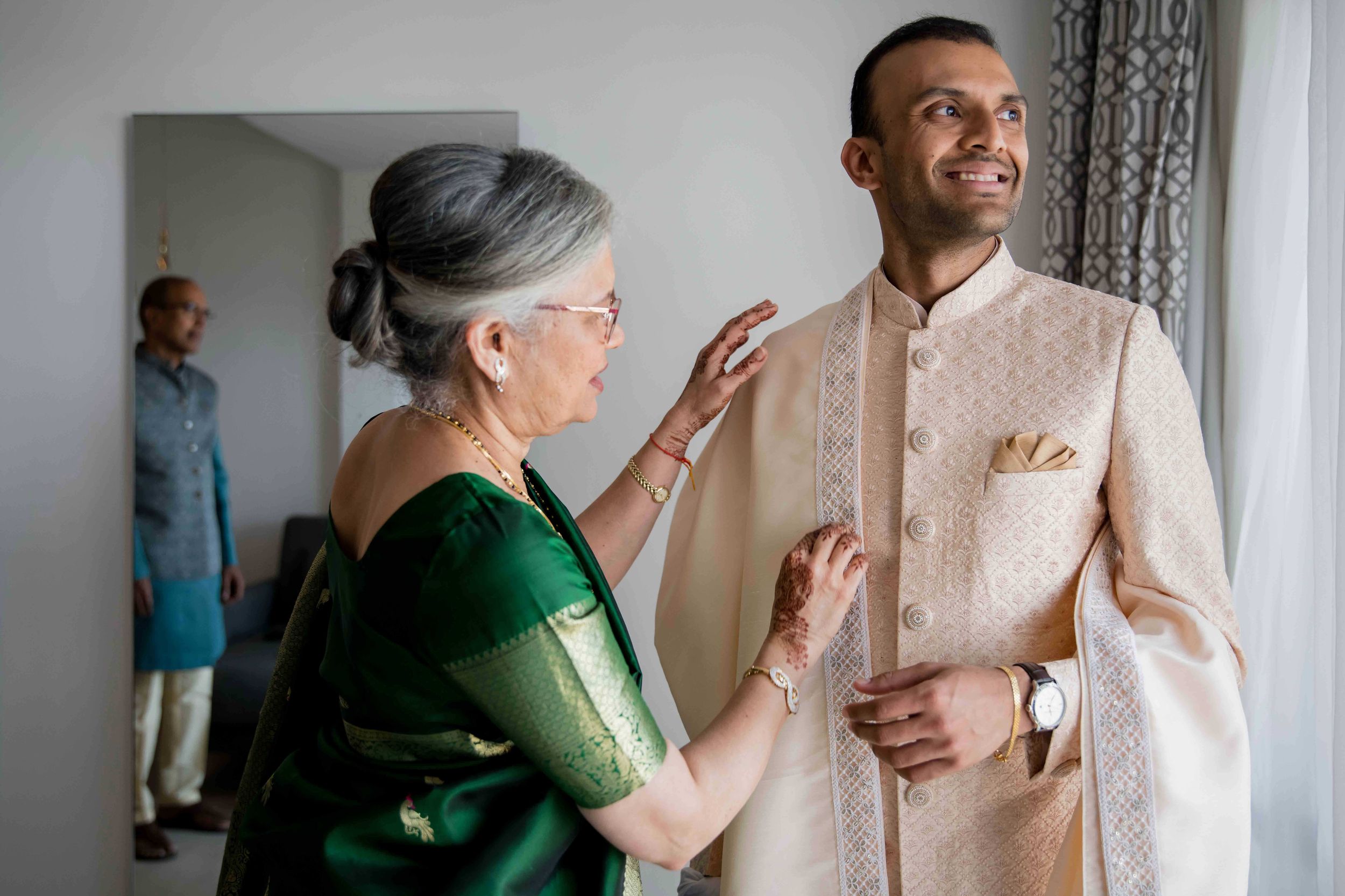 A person in a cream-colored traditional sherwani receives help with their attire from a woman in an emerald green sari.