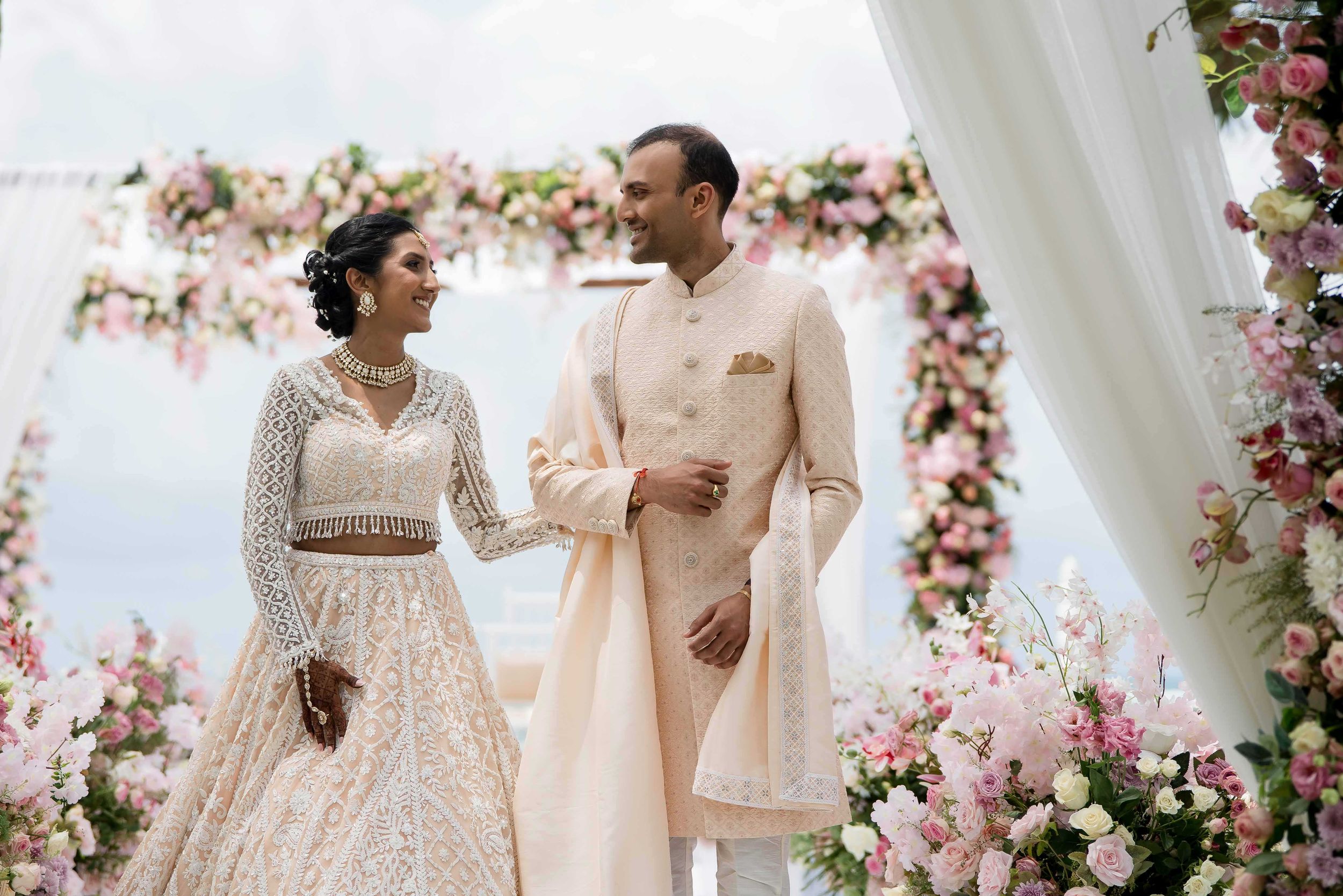 A couple in elegant ivory wedding attire poses amid pink floral decor at their traditional Indian wedding celebration.