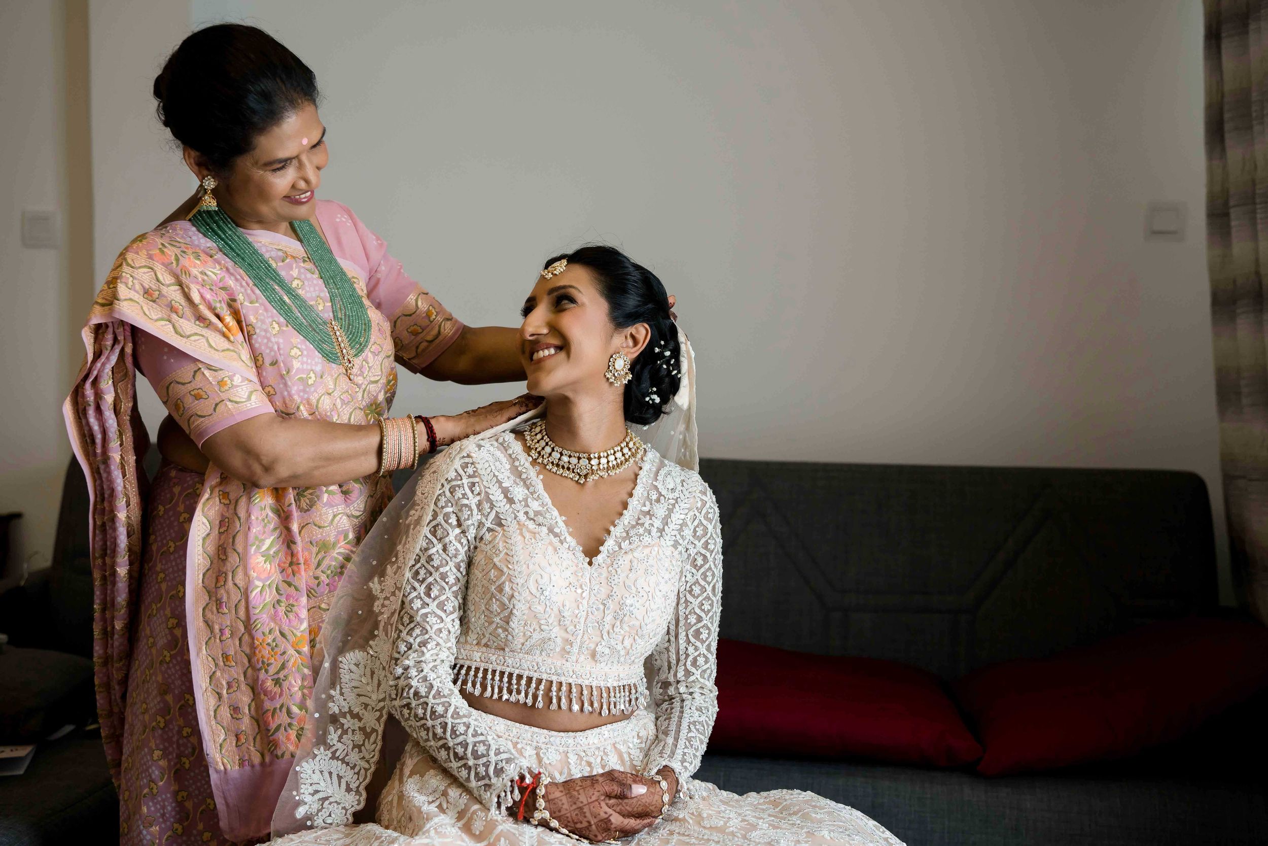 Two people share an intimate moment before a wedding ceremony, with one wearing a pink sari and another in white lace.