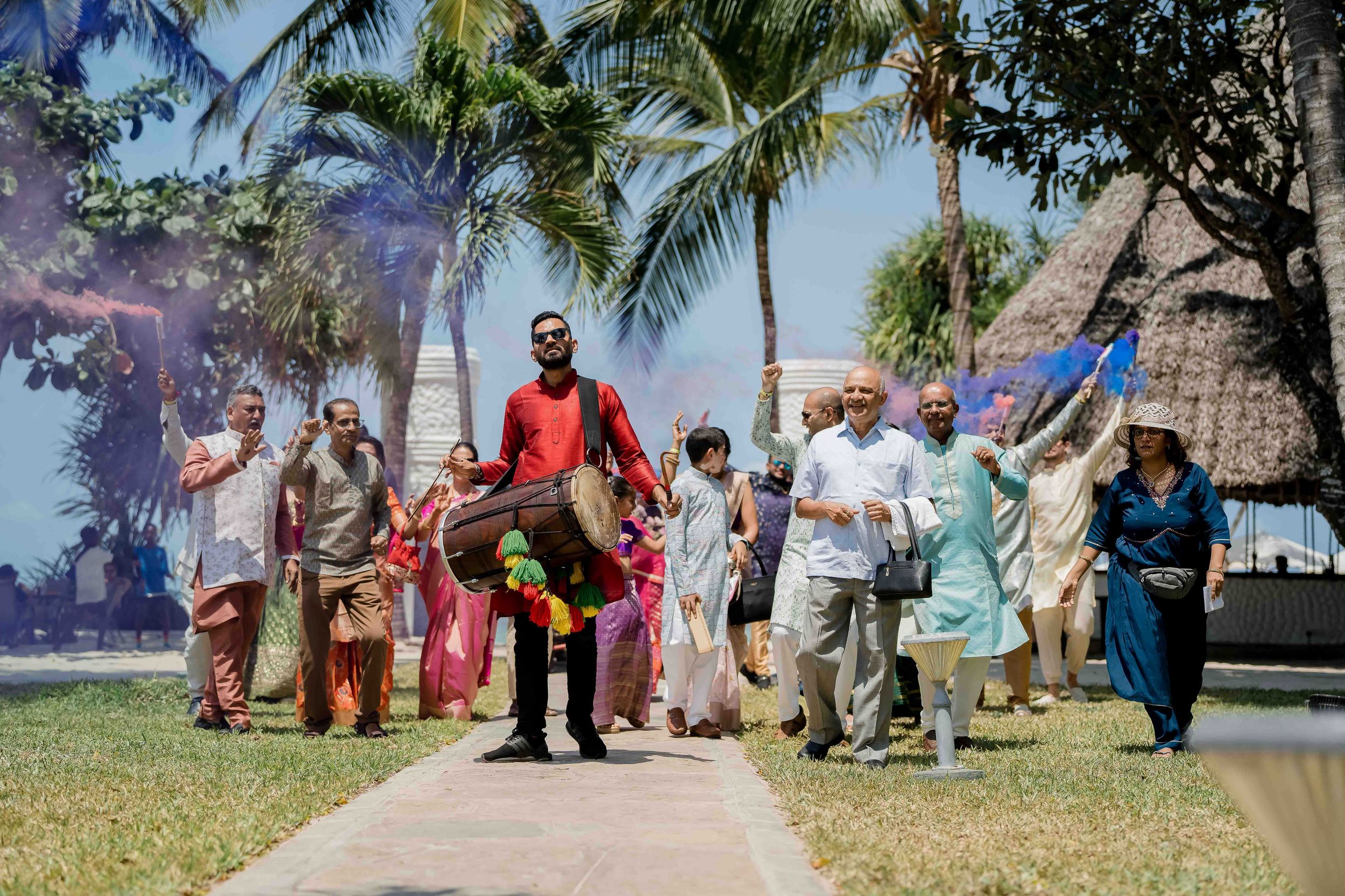 A joyful group celebrates outdoors with traditional attire amidst palm trees and tropical scenery.