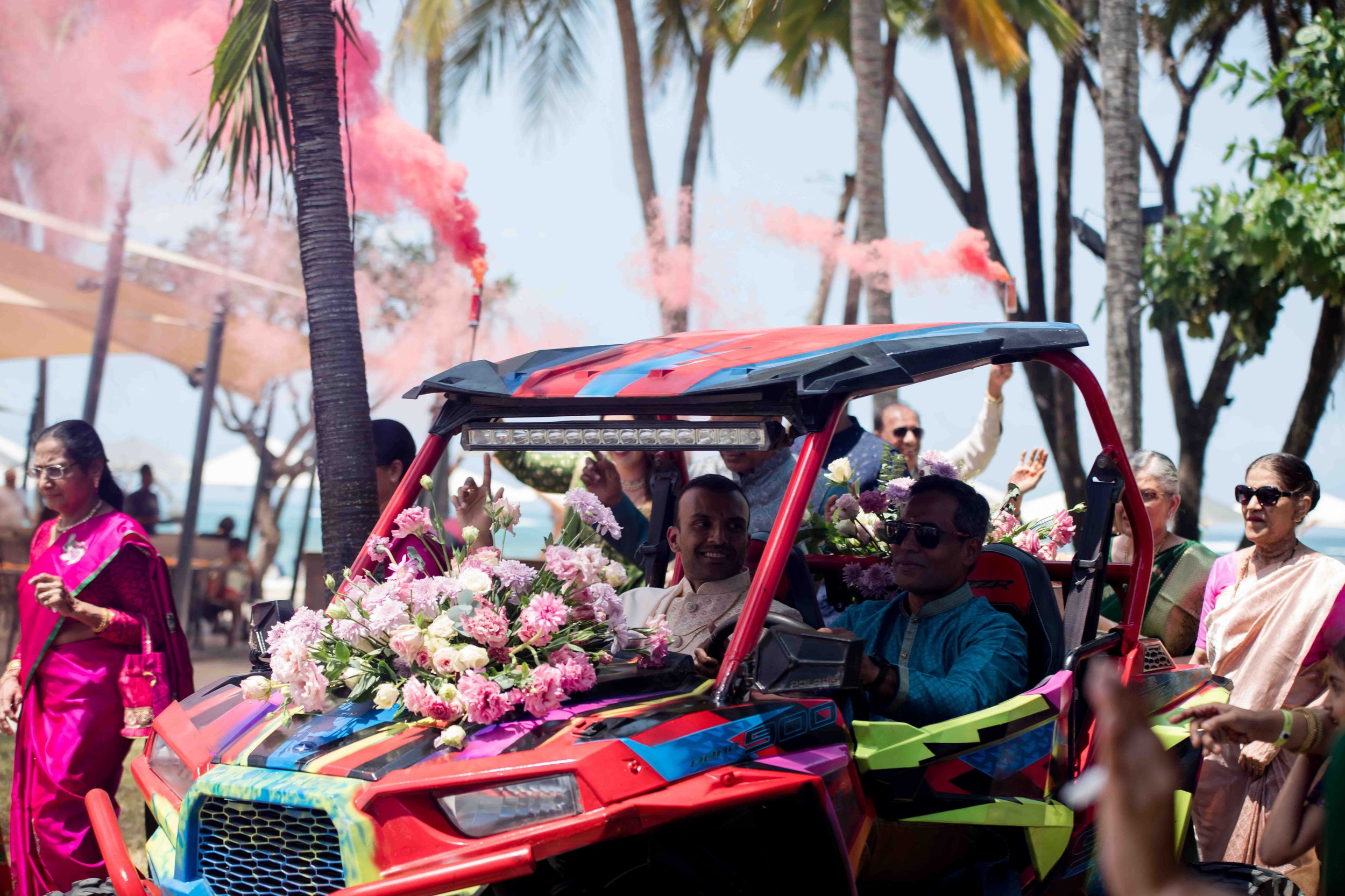 Vibrant outdoor beachside party scene with palm trees, pink smoke and people dancing around bright red sports cars.