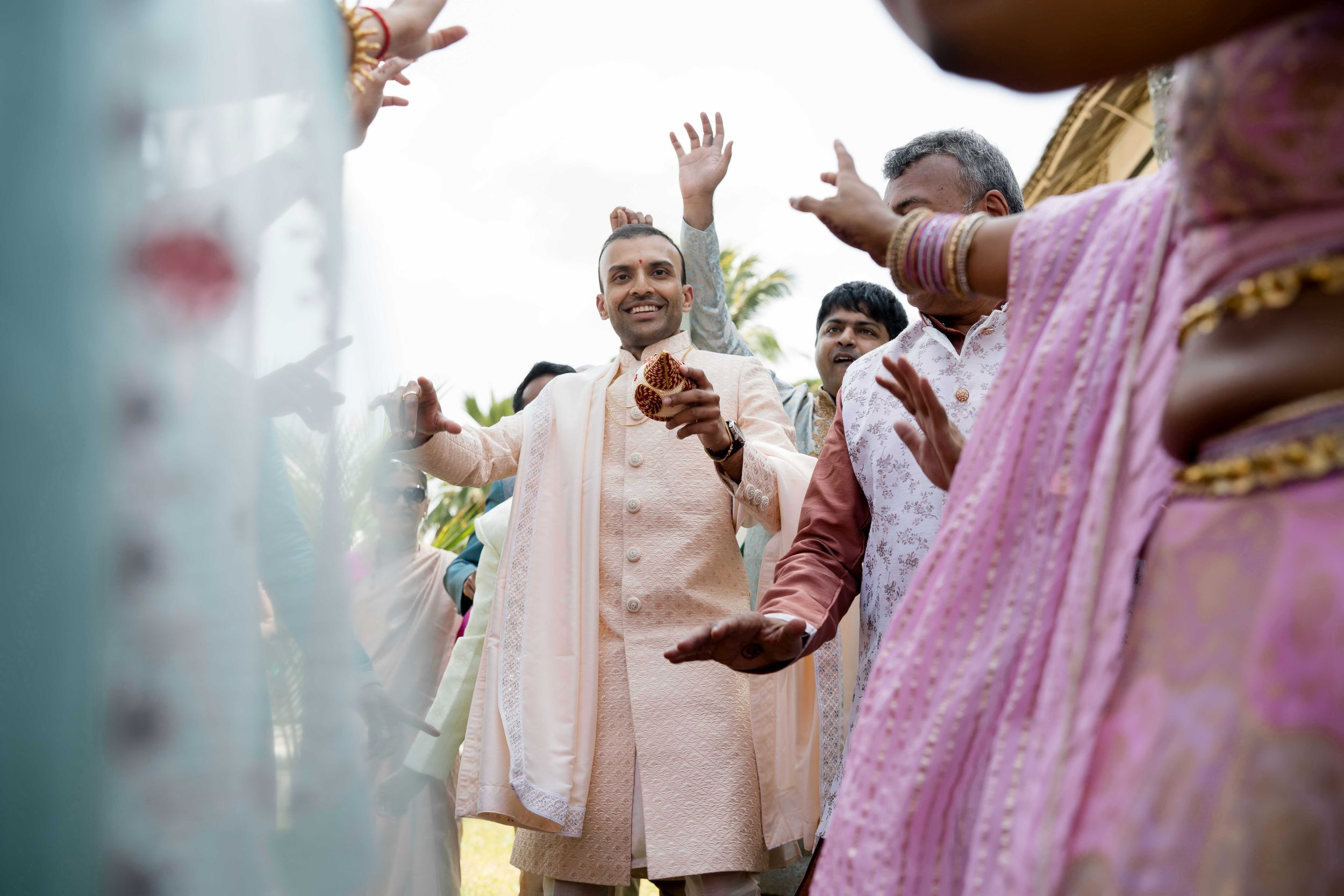 Wedding guests in pastel outfits celebrate with raised hands during a festive outdoor celebration.