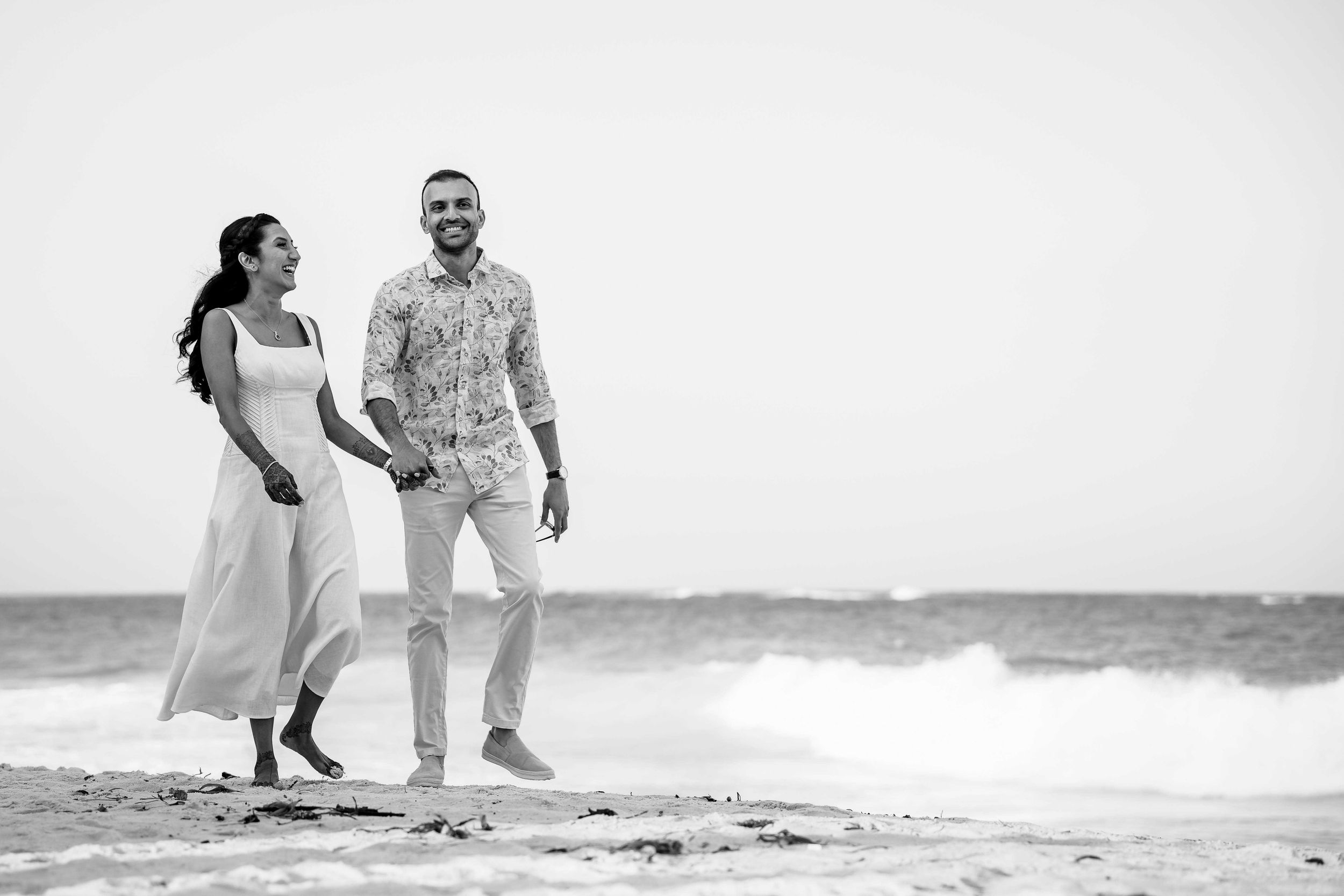 A couple walks hand in hand along a beach shore in black and white photography.