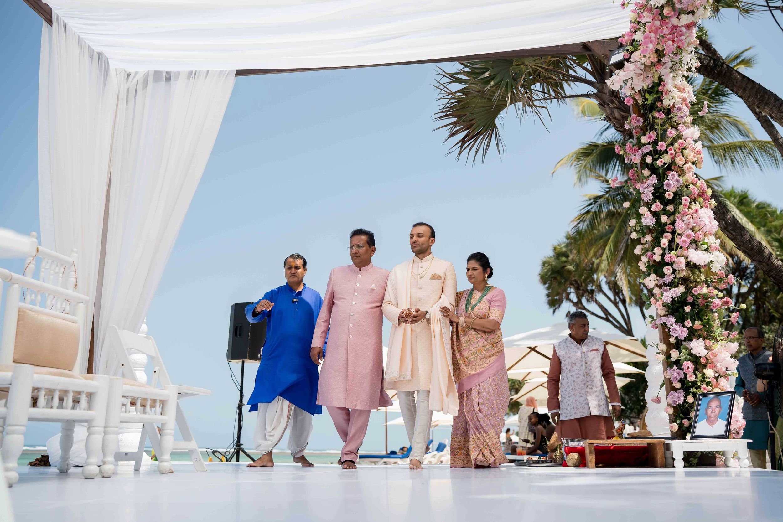 A seaside wedding ceremony setup with white drapes, floral arch, and guests in traditional Indian attire on a sunny day.