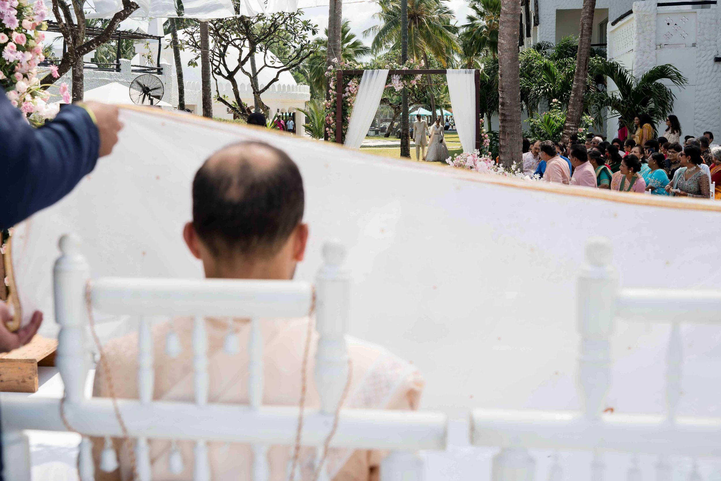 Wedding guests sit in rows of white chairs while tropical palm trees sway in the background during an outdoor ceremony.