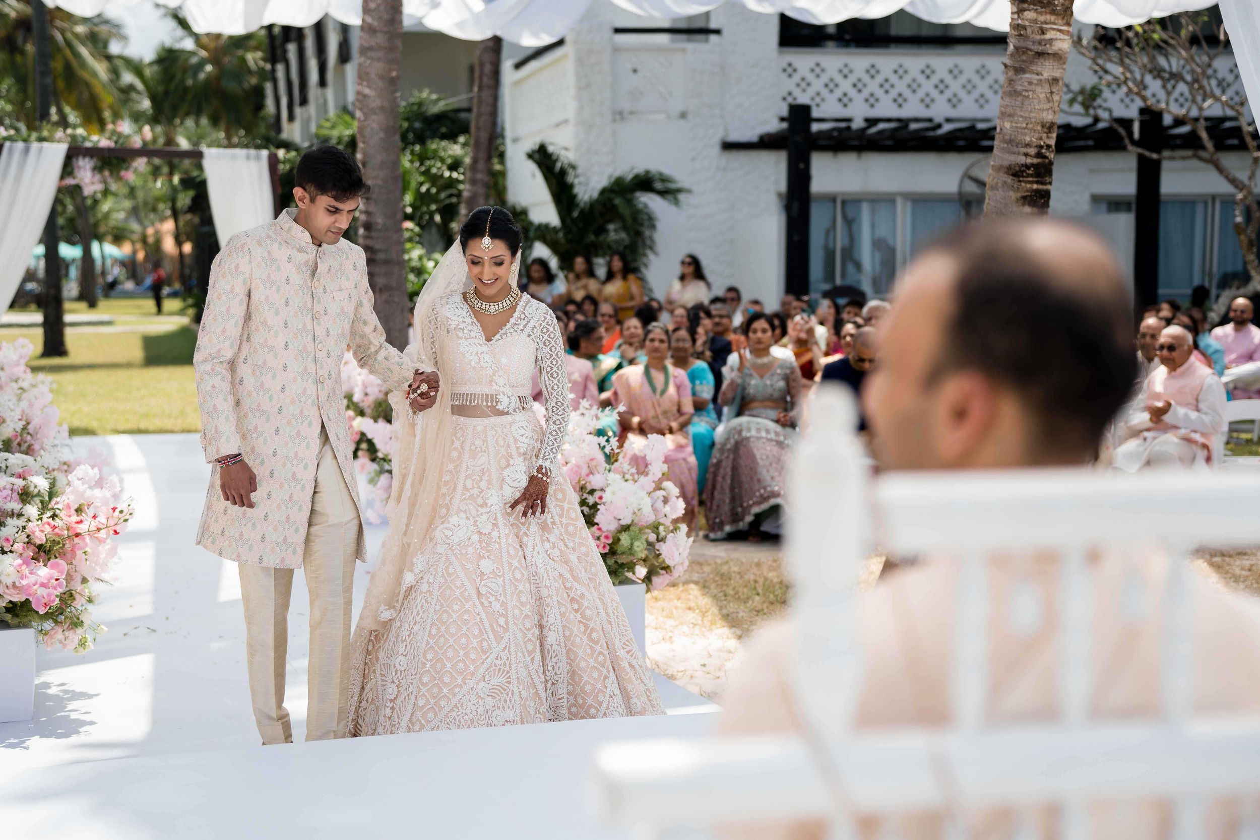 Wedding guests stand on a white aisle runner surrounded by palm trees at an elegant outdoor ceremony.