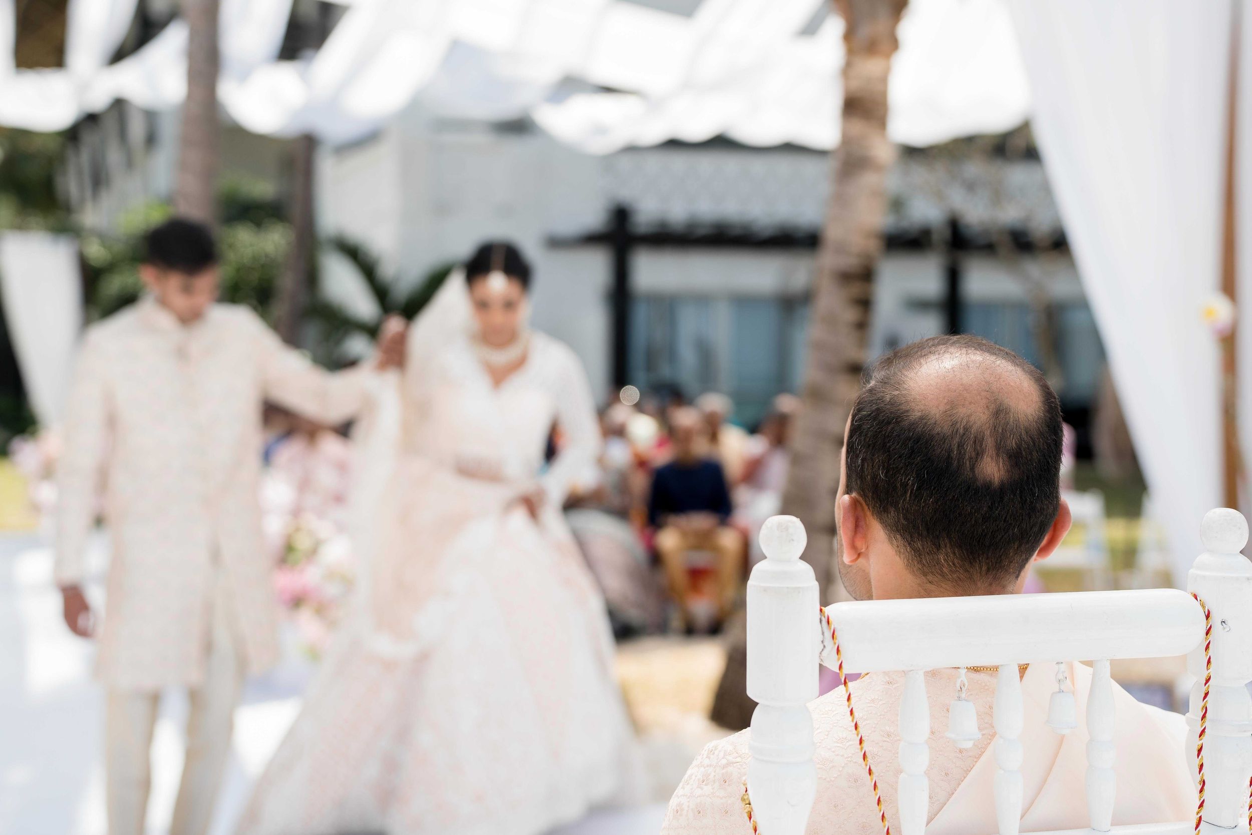 A wedding celebration at an outdoor venue with people dressed in white under palm trees.