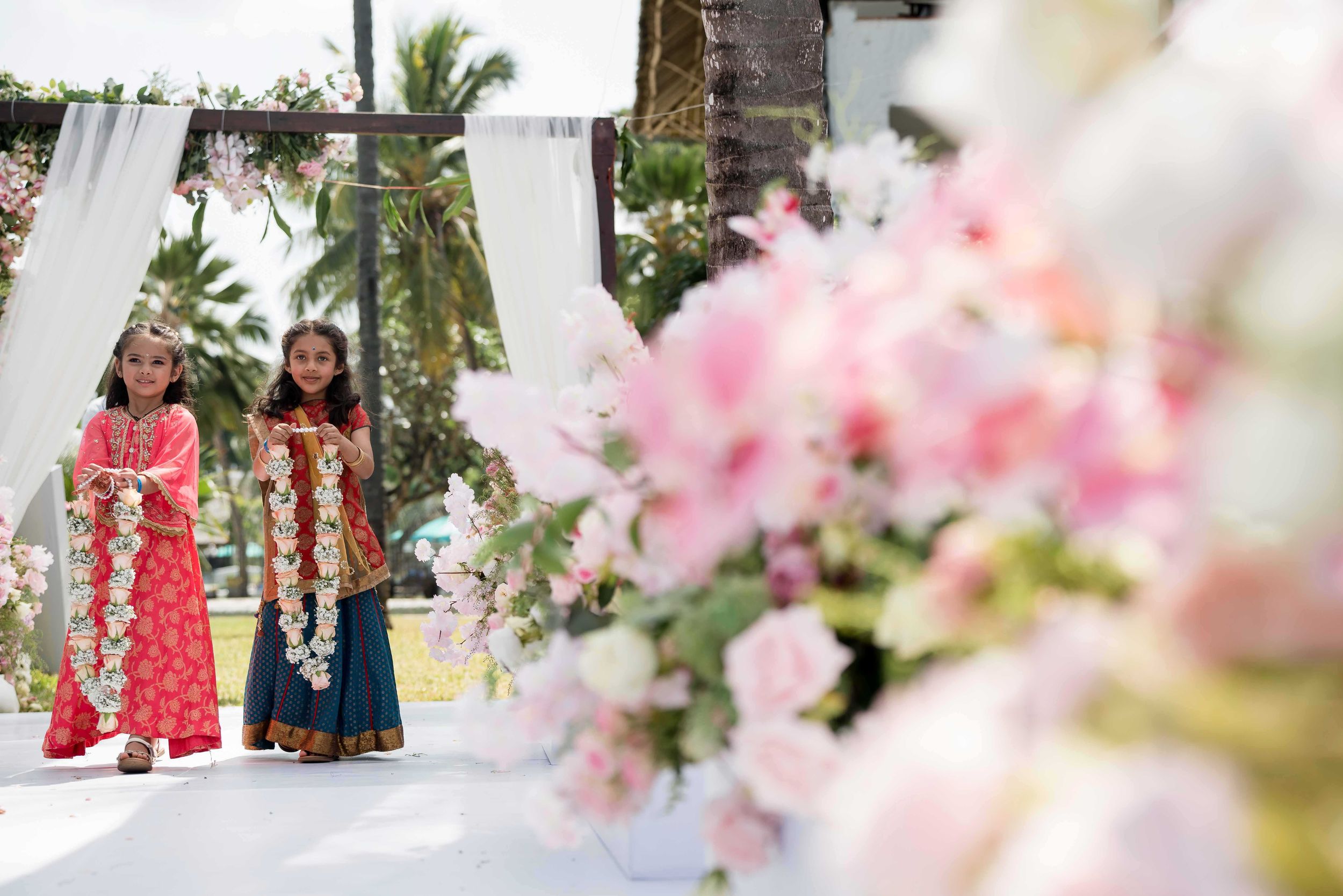 Soft pink floral decorations and white curtains create an elegant outdoor wedding ceremony setup with palm trees in background.