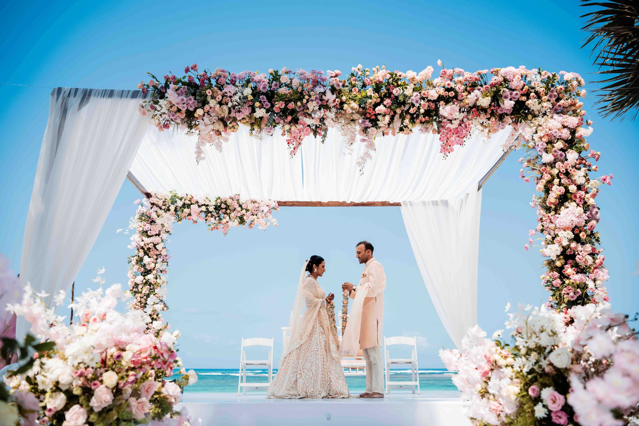 A wedding ceremony under a floral arch draped with white fabric overlooking the ocean on a bright sunny day.