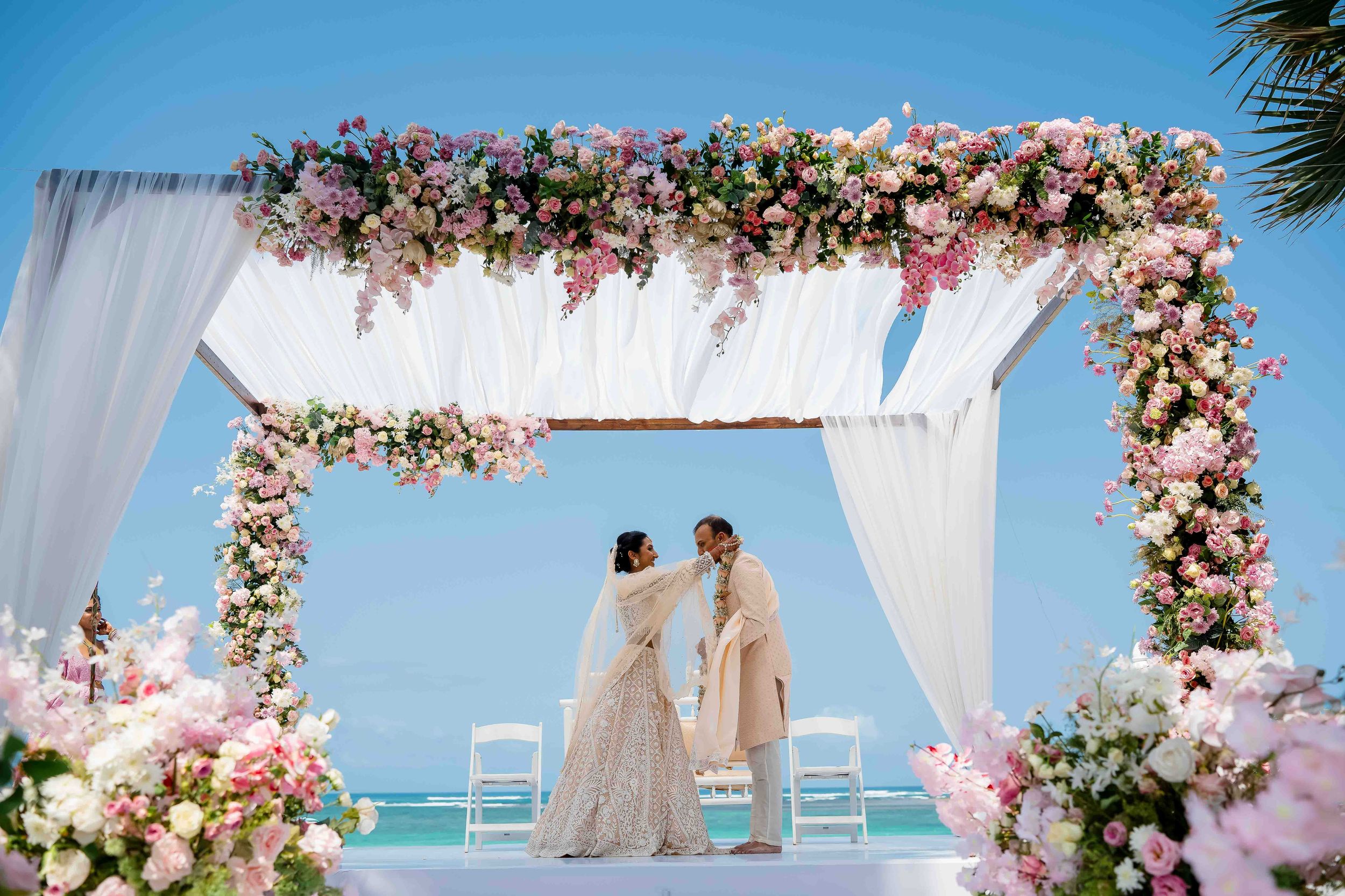 Wedding ceremony setup with white draped canopy adorned with pink and white flowers on a beachfront overlooking the ocean.