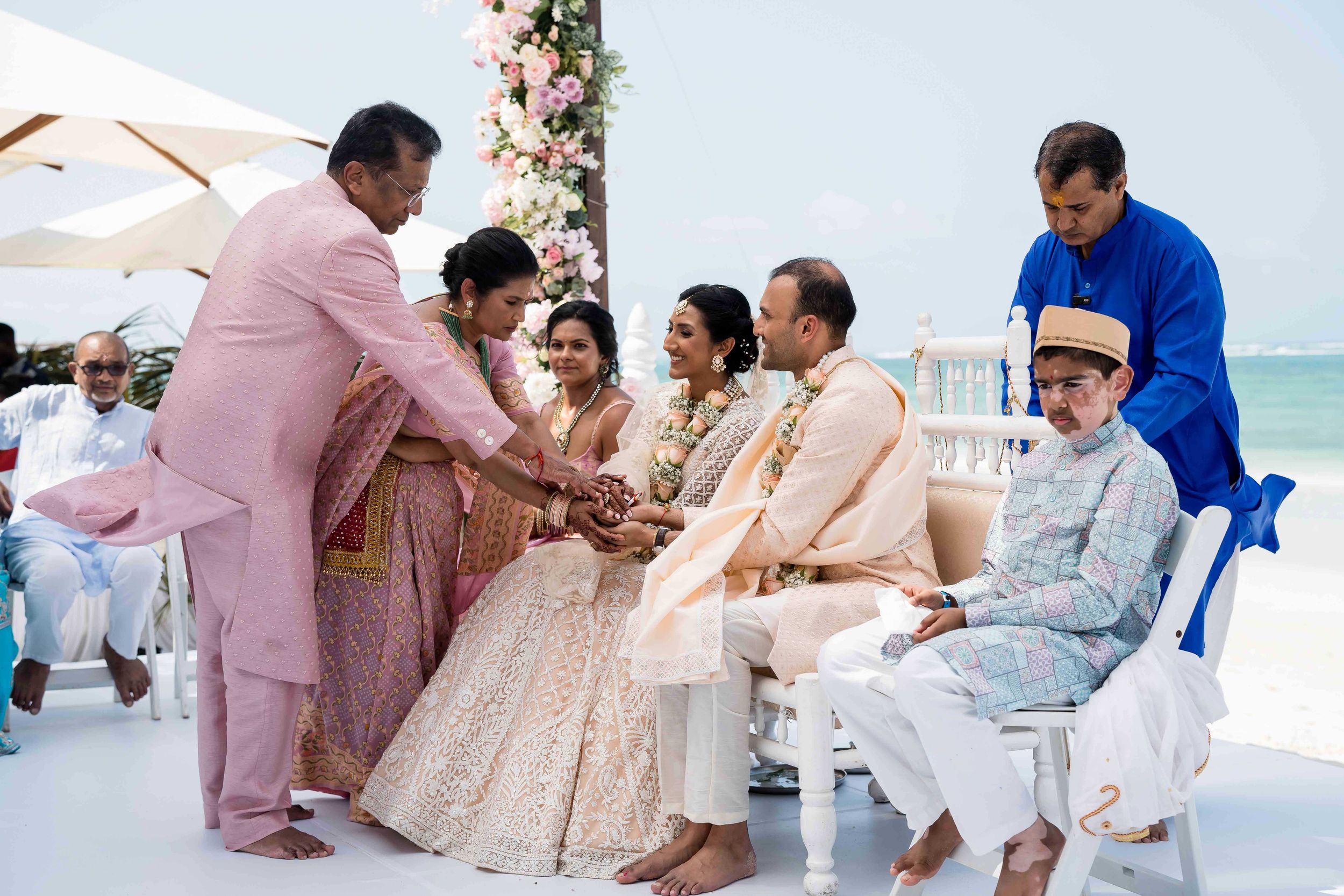 A traditional Indian wedding ceremony takes place on a white platform by the ocean with guests in pastel formal attire.