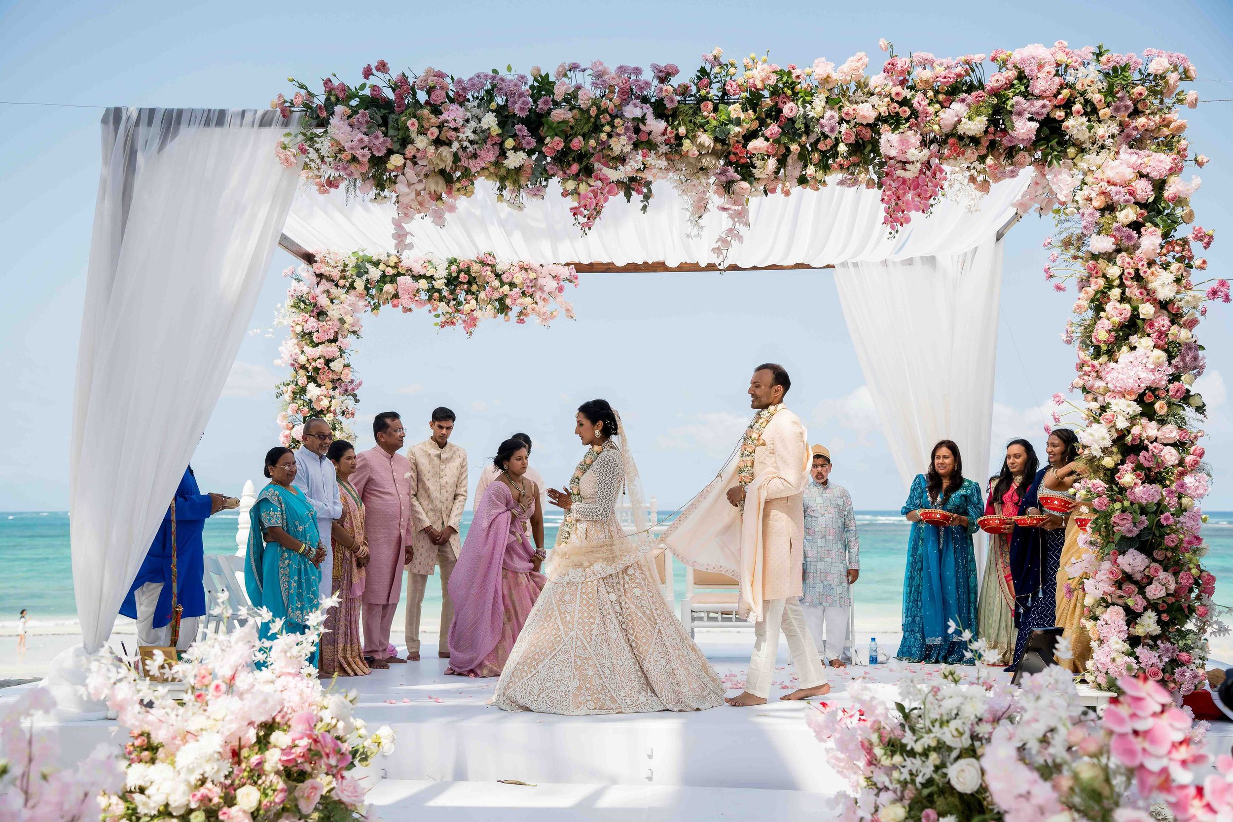 A dreamy beach wedding ceremony setup with pink and white floral arches and an ocean view backdrop at sunset.