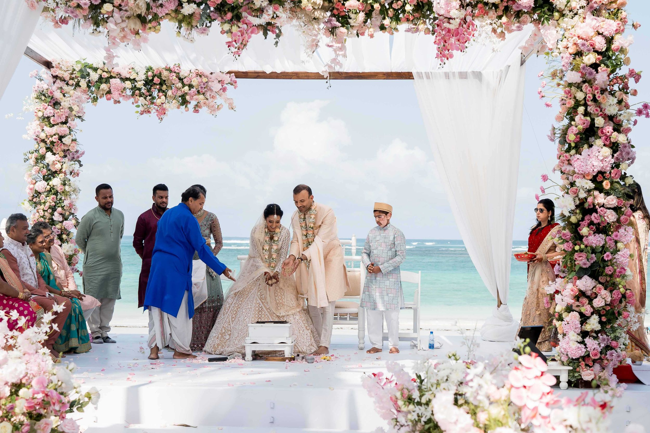 Beachfront wedding ceremony setup with white canopy and pink floral arrangements alongside turquoise ocean waters.
