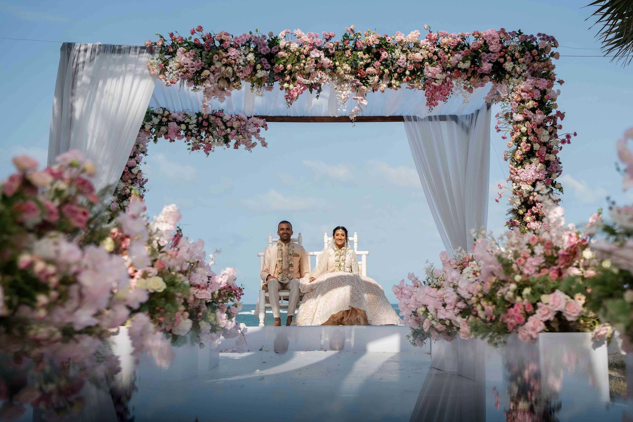 Outdoor wedding ceremony setup with white drapes and pink floral arches against a blue sky backdrop.