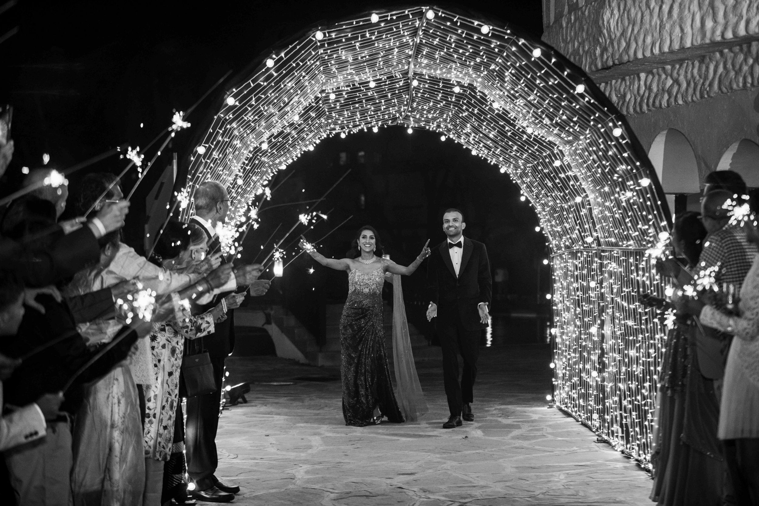 Wedding party exits venue through a magical light tunnel archway as guests celebrate in black and white.