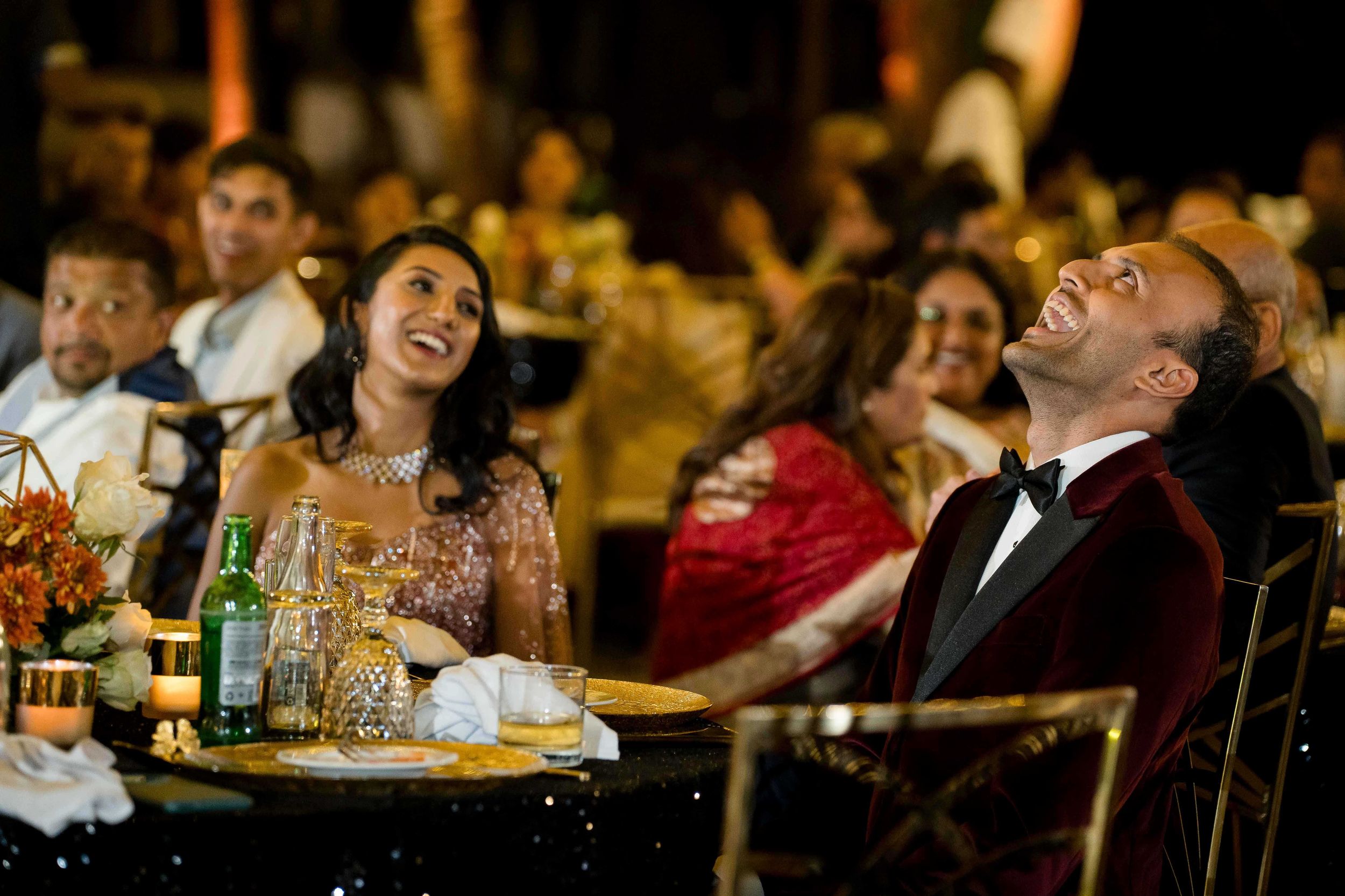 Attendees at a formal dinner event laugh heartily while seated at tables with drinks and glassware.