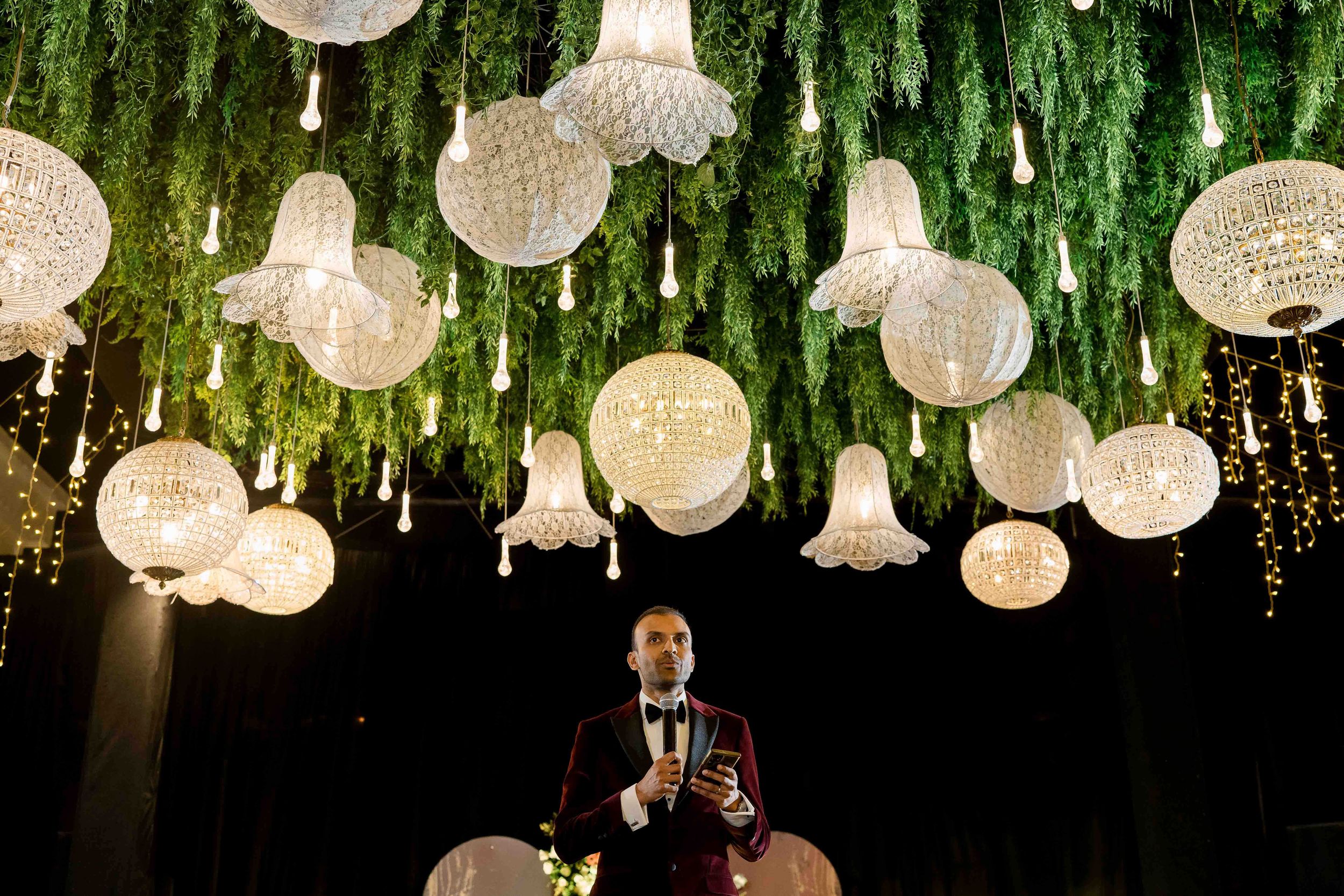 Crystal chandeliers and orb-shaped pendant lights hang elegantly from a dark ceiling with cascading greenery decorations.