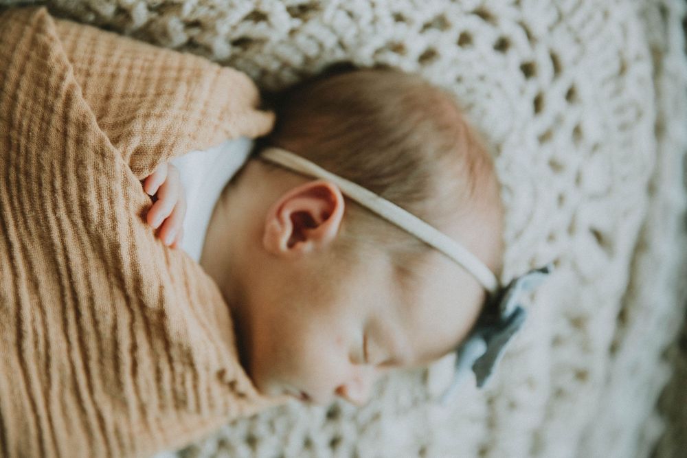A sleeping infant swaddled in a soft peach blanket rests peacefully with a white band across their forehead.