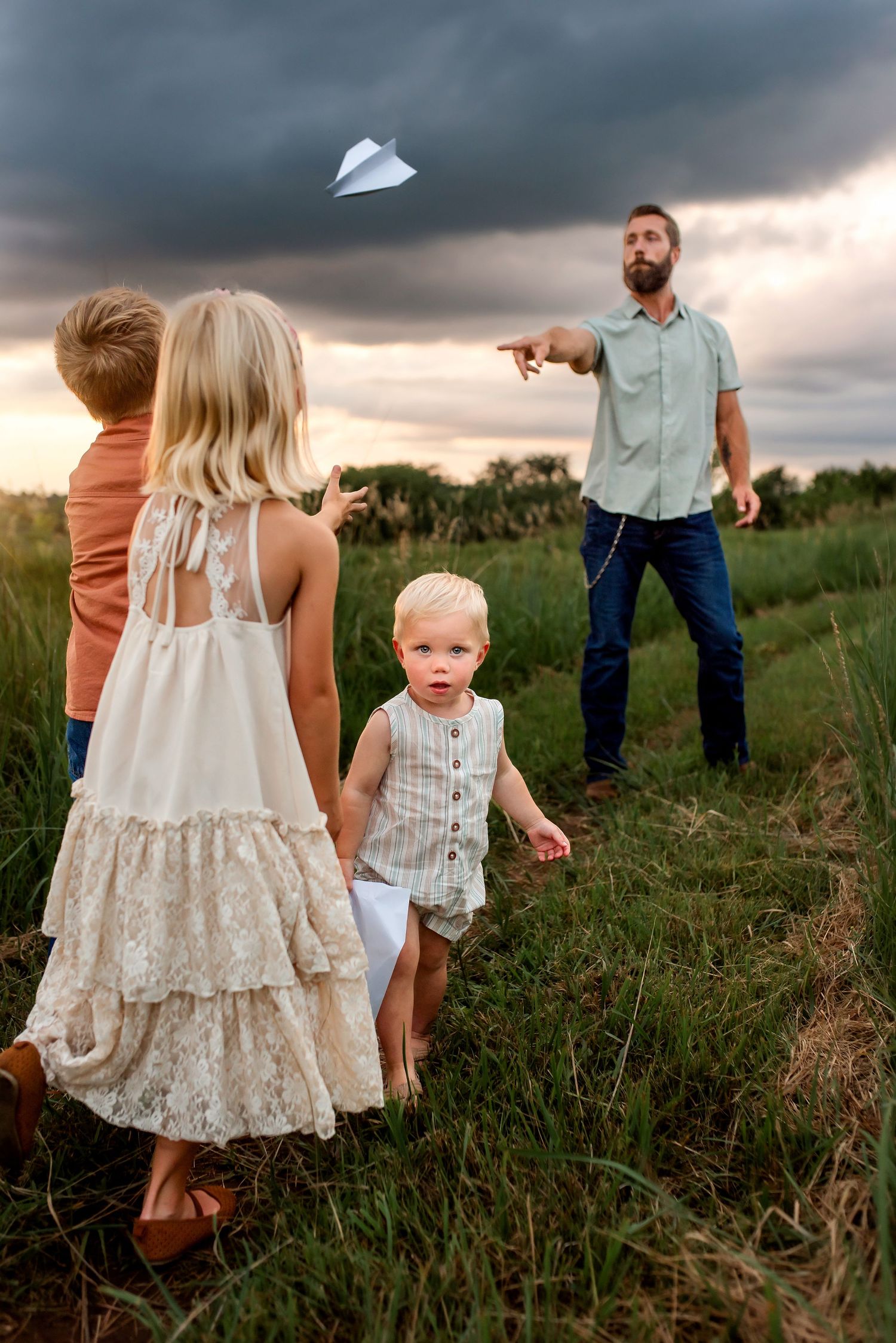 Family playing with paper airplane in grassy field during stormy sunset with dramatic clouds overhead.
