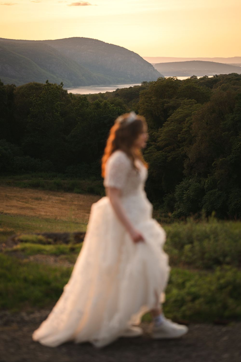 Figure in flowing white wedding dress walks along hilltop path at sunset with mountains and lake in background.