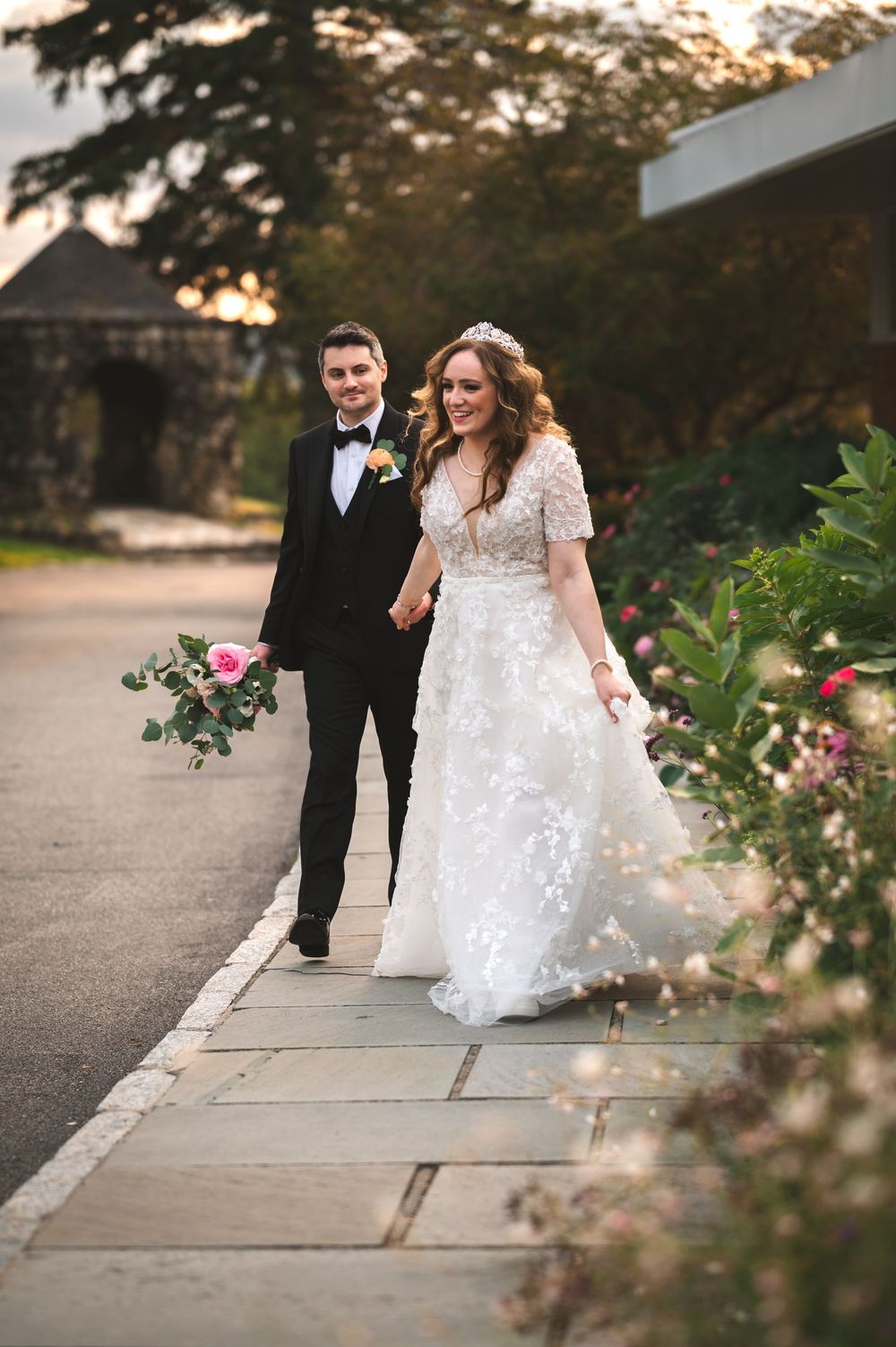 A couple in formal wedding attire walk together along an outdoor pathway framed by flowering rose bushes at sunset.