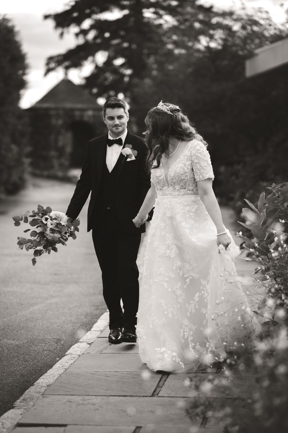 A bride and groom walk together on a garden path, captured in elegant black and white wedding photography.