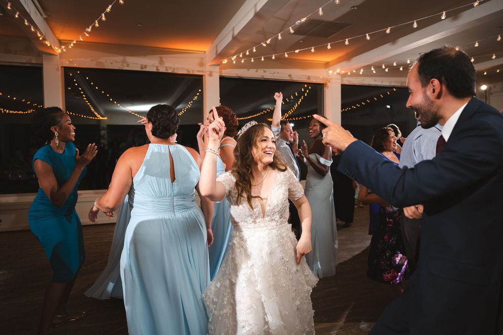 People dancing and celebrating at an indoor wedding reception under string lights and white ceiling decorations.