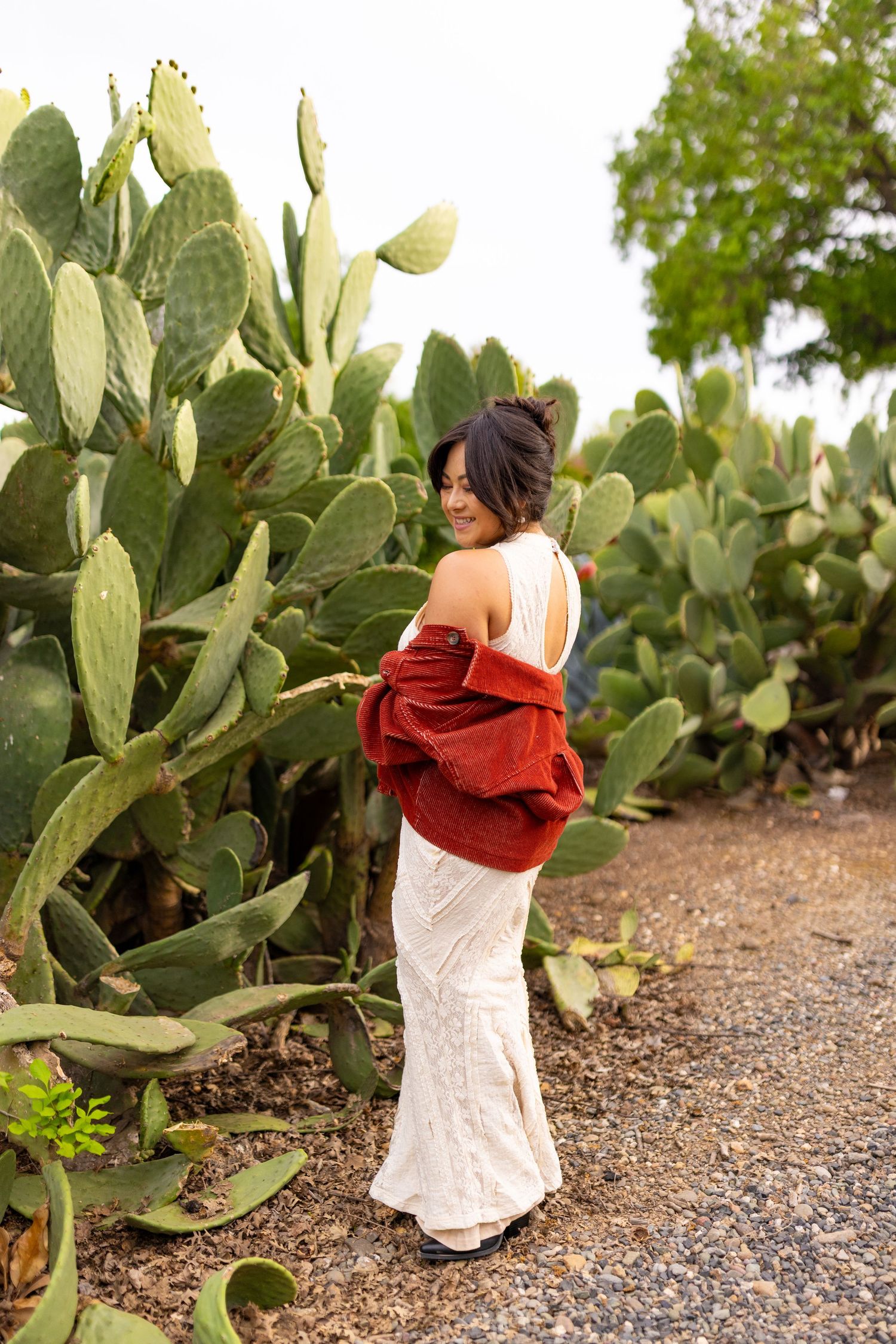 A person in a white dress and red sweater poses beside large prickly pear cactus plants.